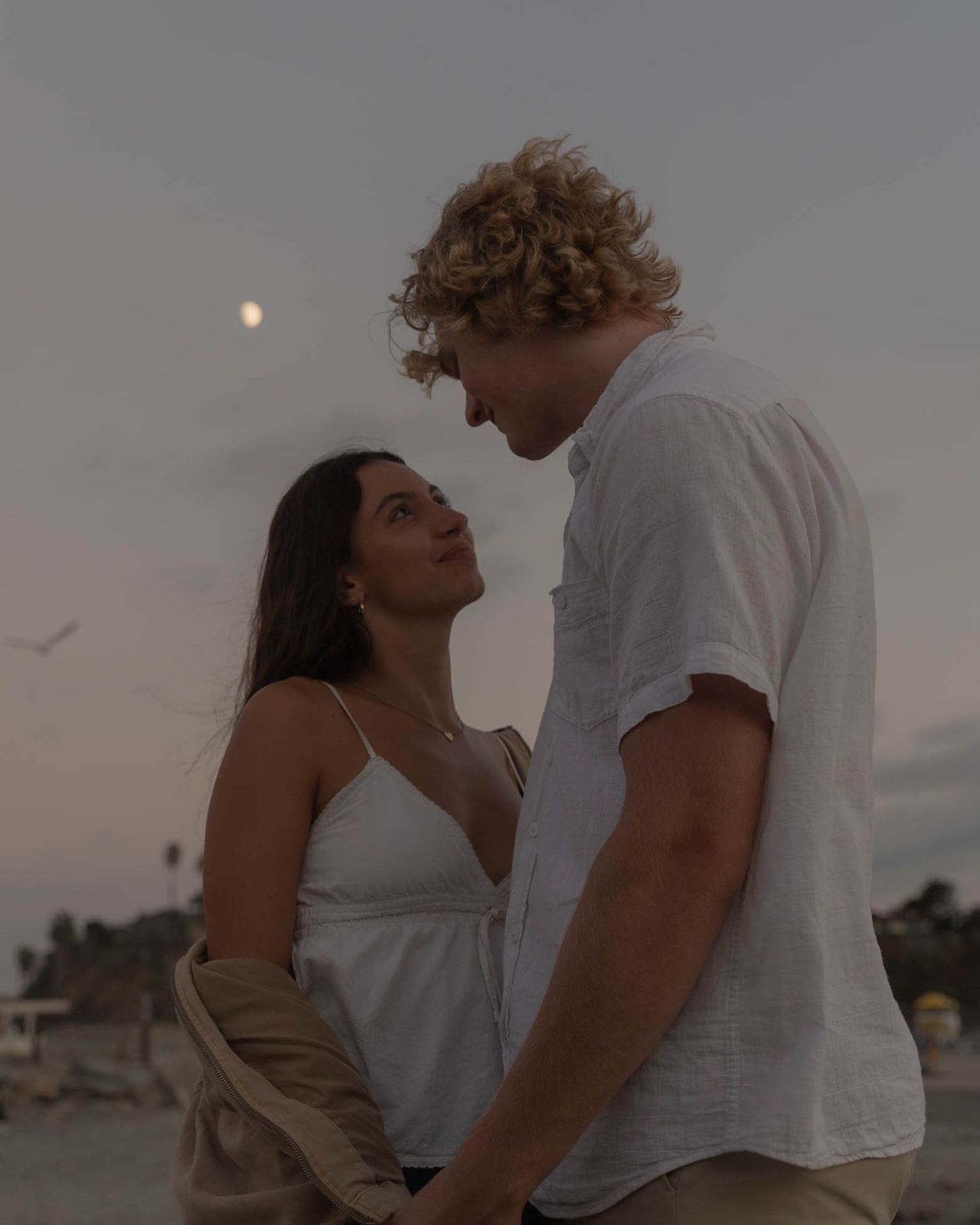Romantic couple embracing on beach at sunset with moon visible in background.
