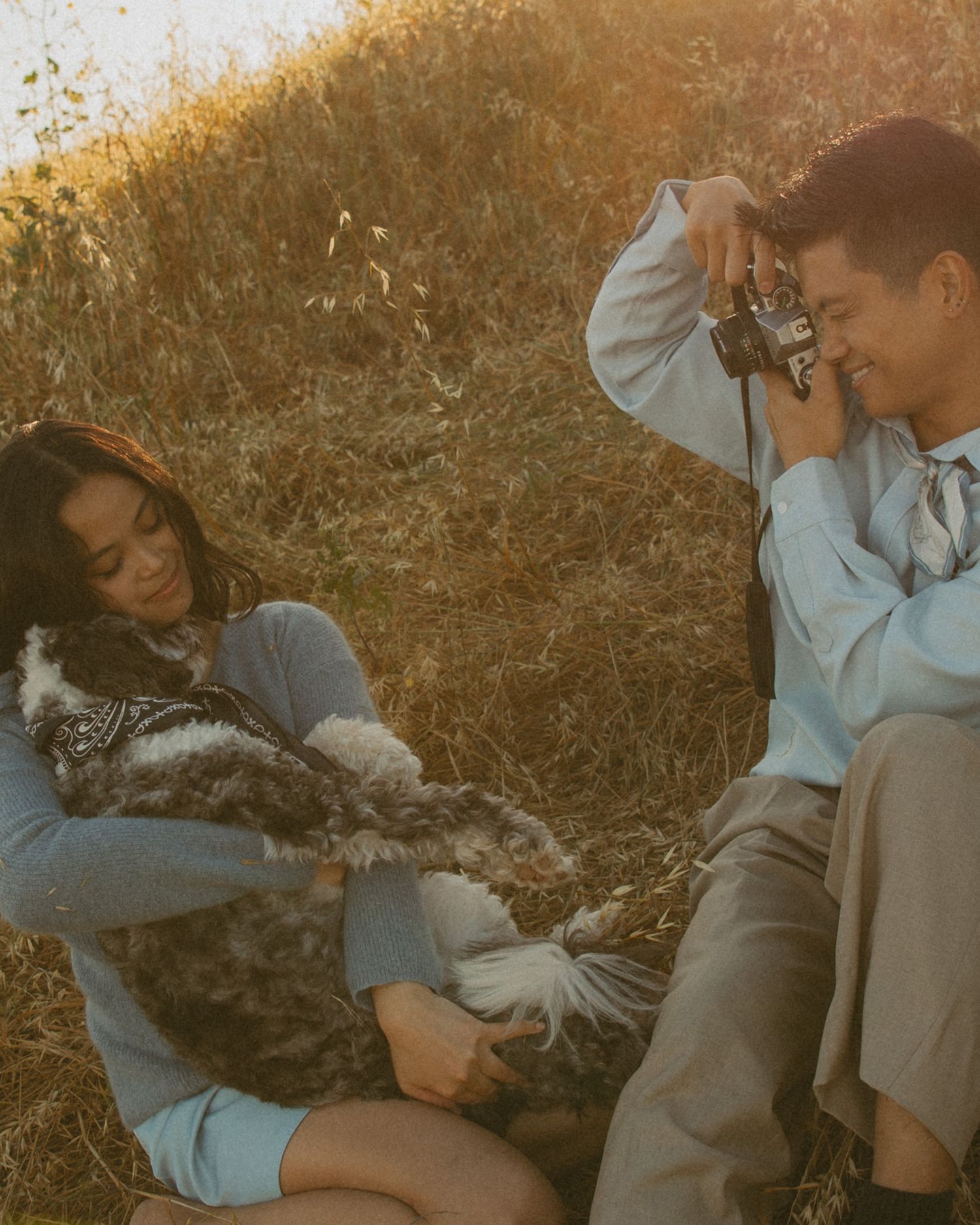 Couple sitting in golden field, woman holding fluffy dog while man photographs with vintage camera at sunset.