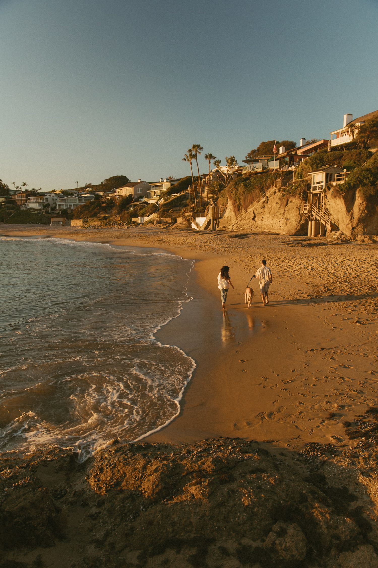 Couple walking on a sandy beach at golden hour with cliffside homes and palm trees in the background.