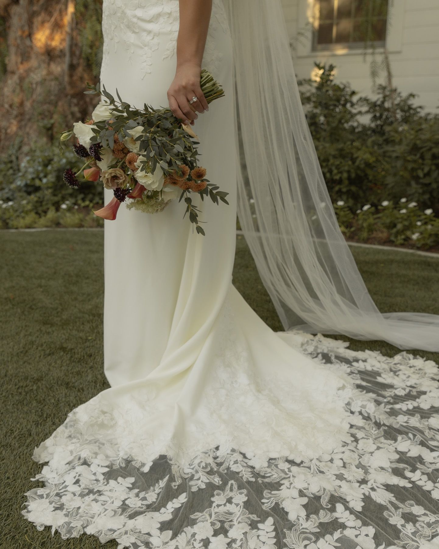 Bride in elegant lace-trimmed white gown with cathedral veil holds a rustic fall wedding bouquet.