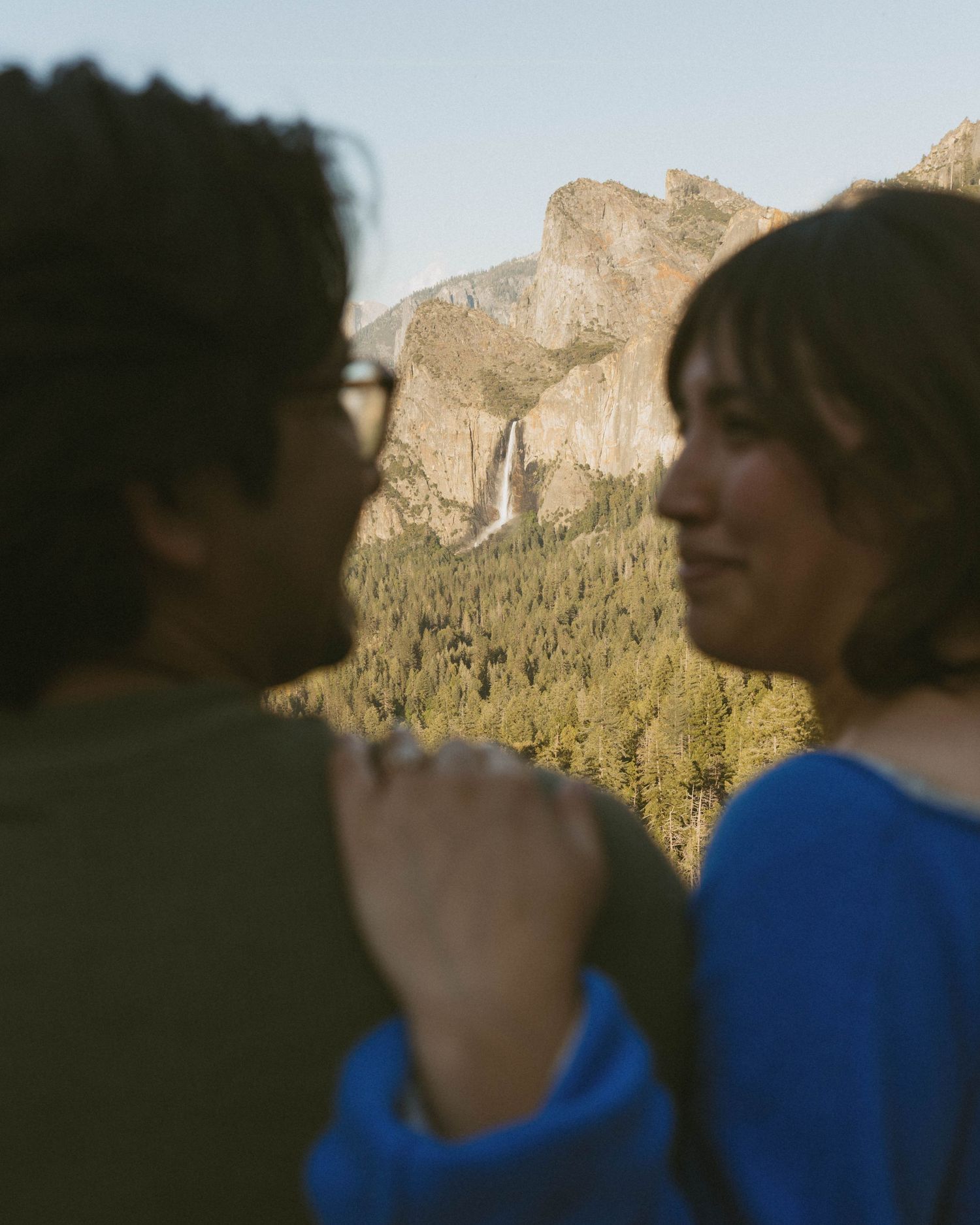 Couple holding hands facing each other with Bridalveil Fall and Yosemite Valley mountains in the background.
