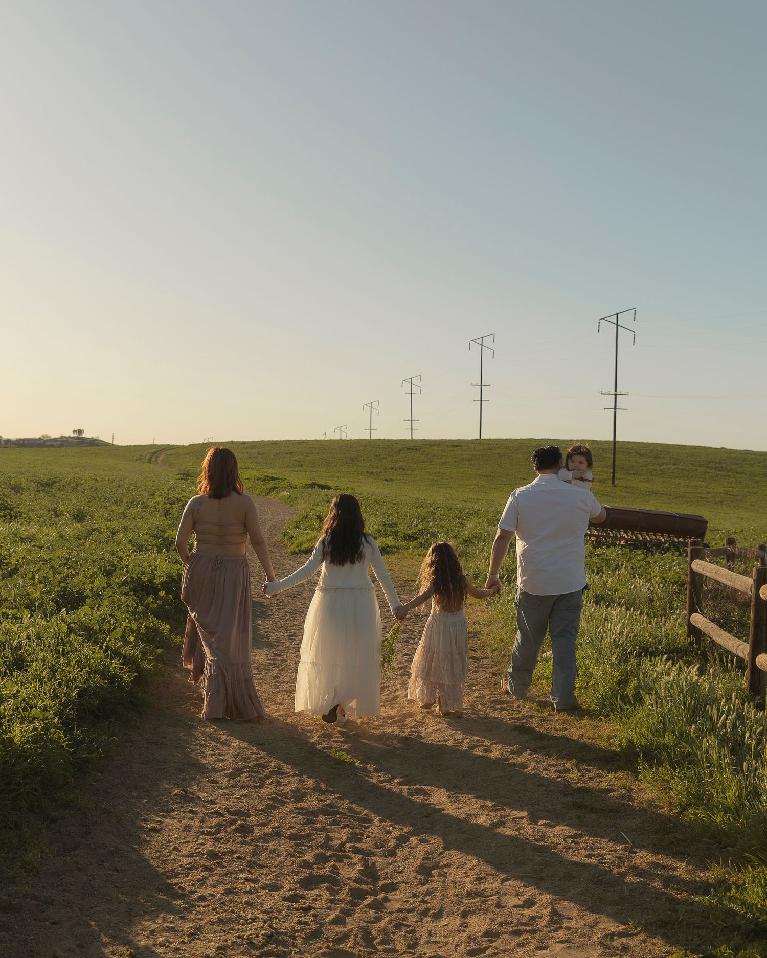 Family of four holding hands walking down a sandy path through green fields at golden hour sunset.