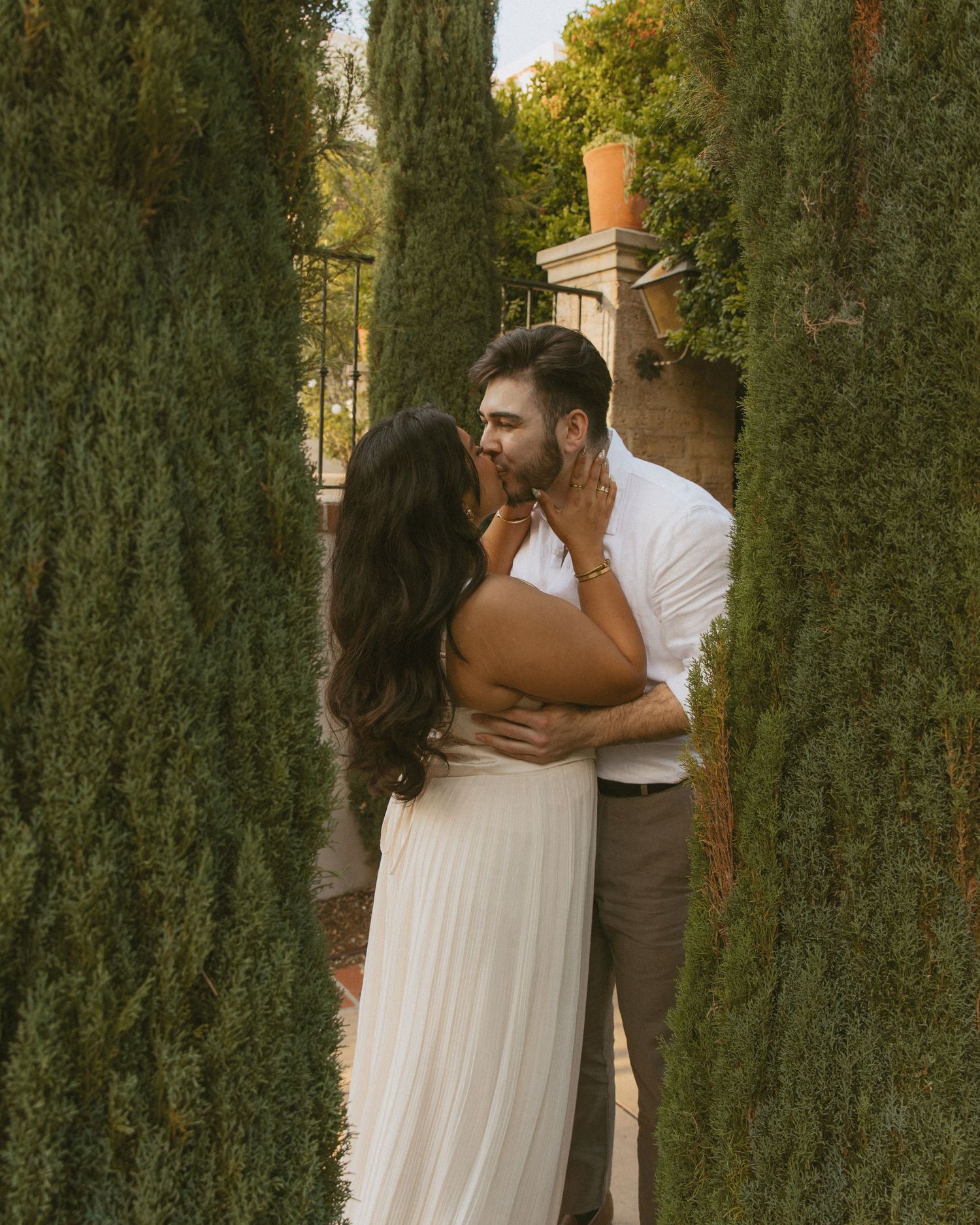A couple shares a romantic kiss among tall cypress trees, the woman in a white dress and man in a white shirt.