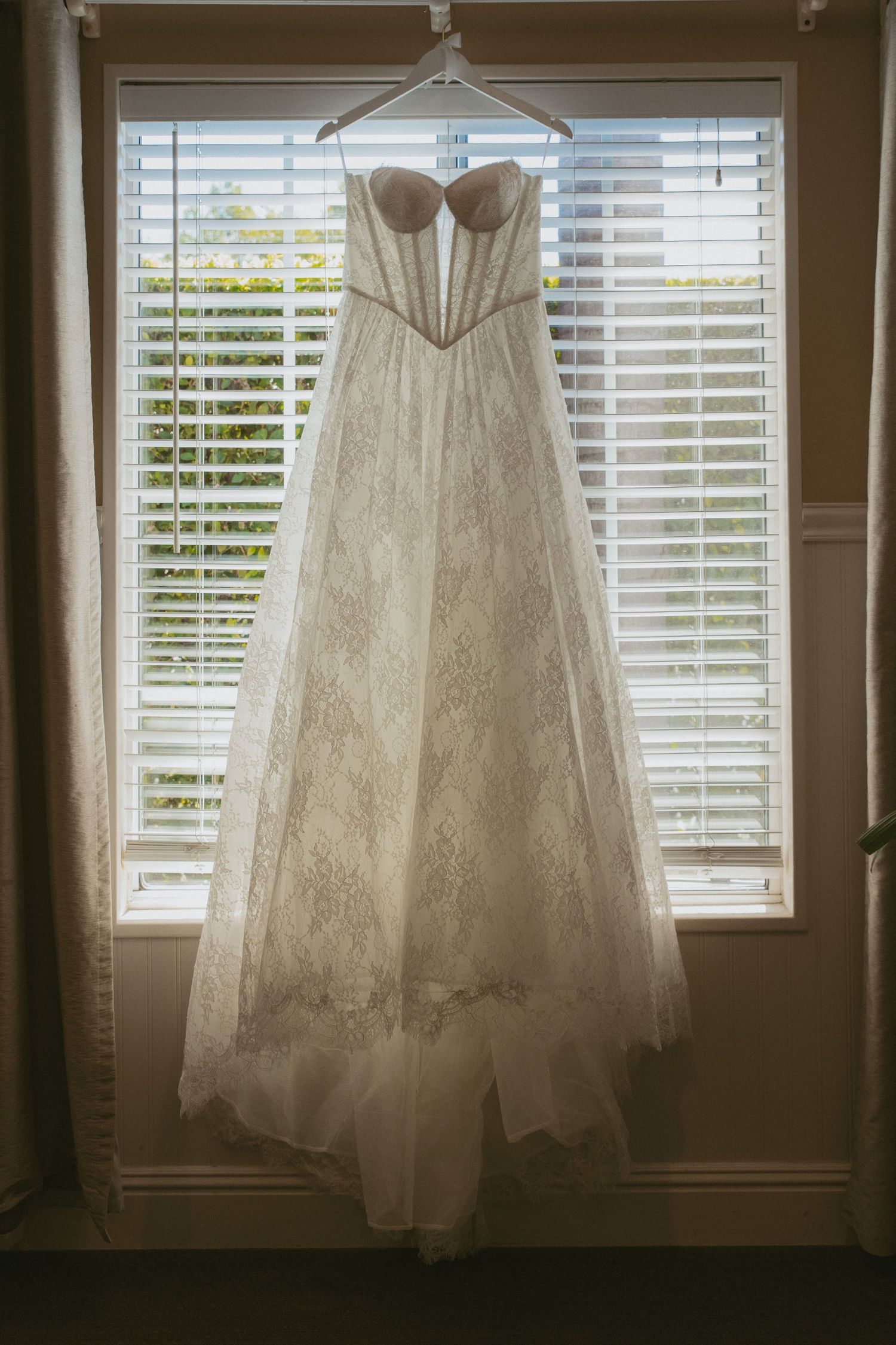 Elegant strapless lace wedding gown hanging on a hanger in front of a sunlit window with white blinds.