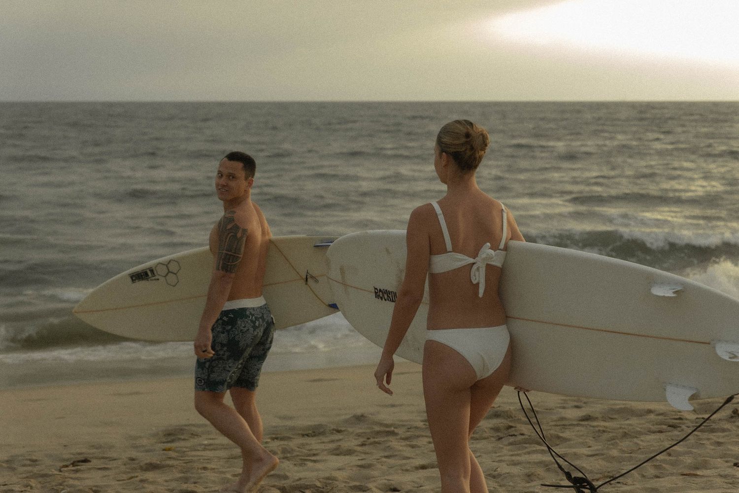 Two surfers carrying their boards along the beach at sunset.