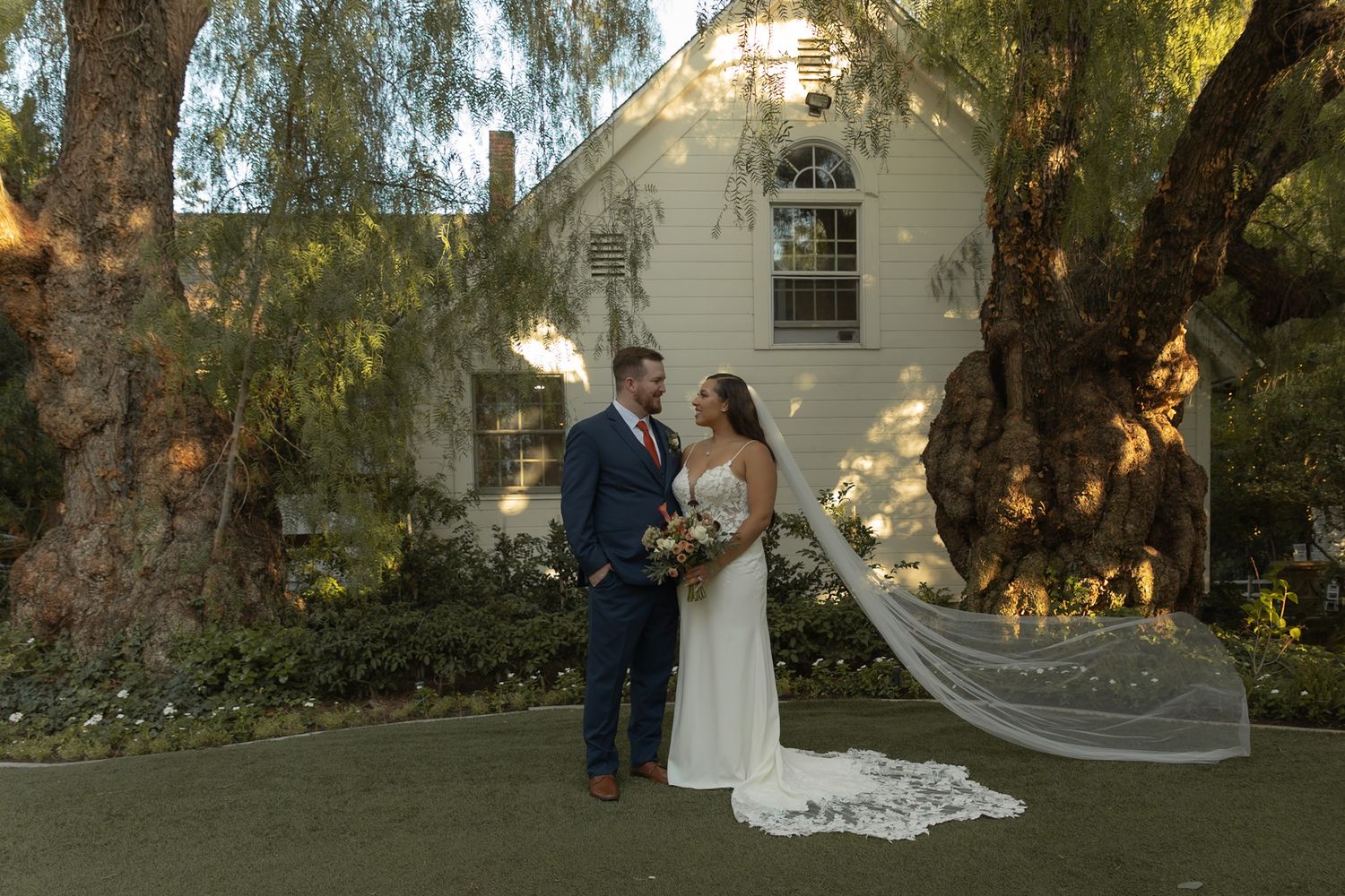 Bride and groom stand together on lush green lawn before a charming white farmhouse at golden hour.