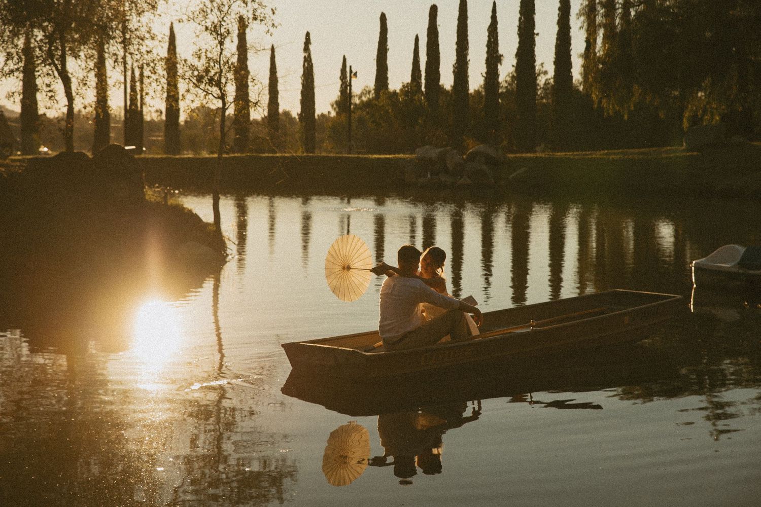 Couple sitting in a wooden rowboat on a serene lake at golden sunset, surrounded by cypress trees.