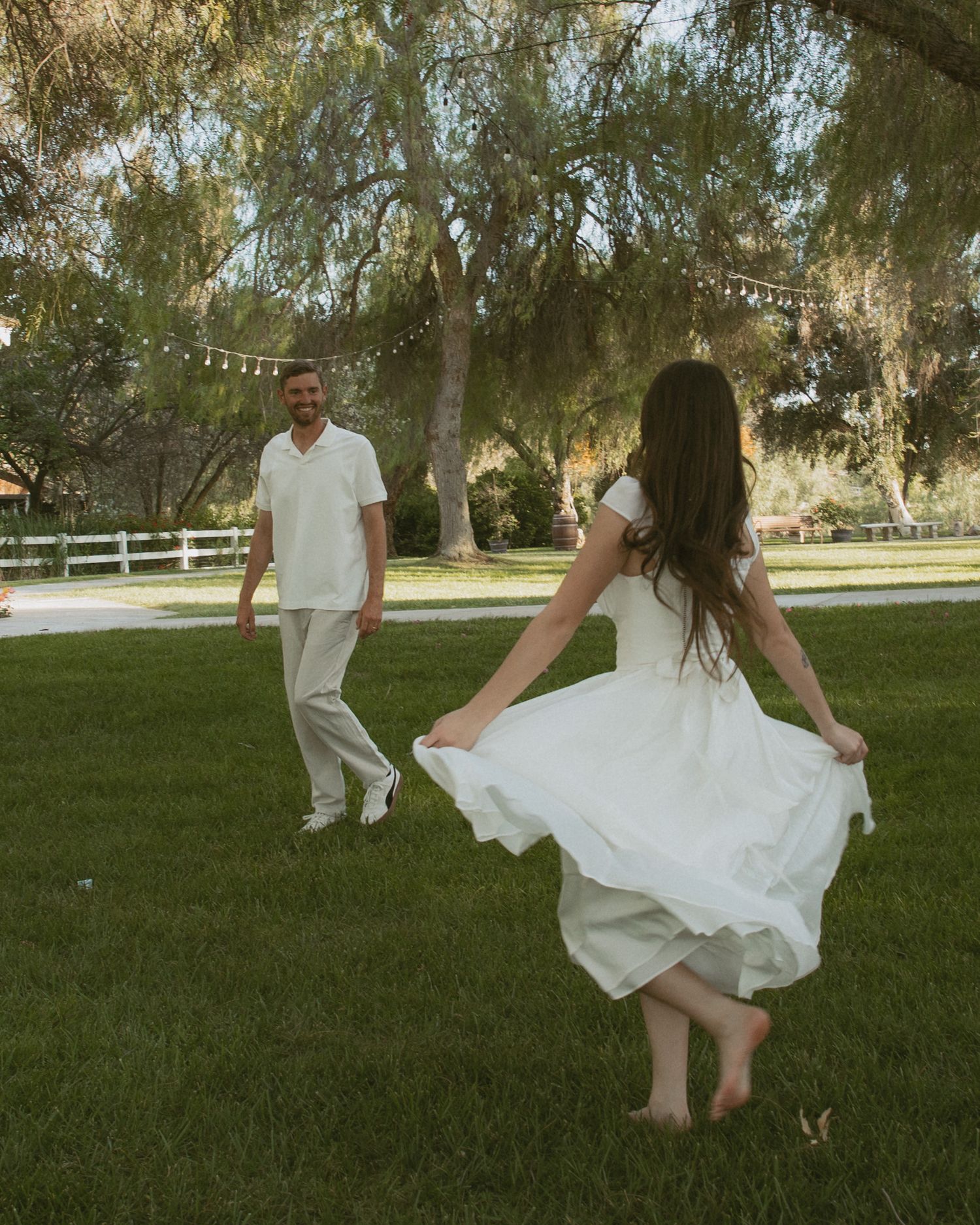 Bride in white dress twirling barefoot on green lawn while groom in white outfit watches under shady trees.