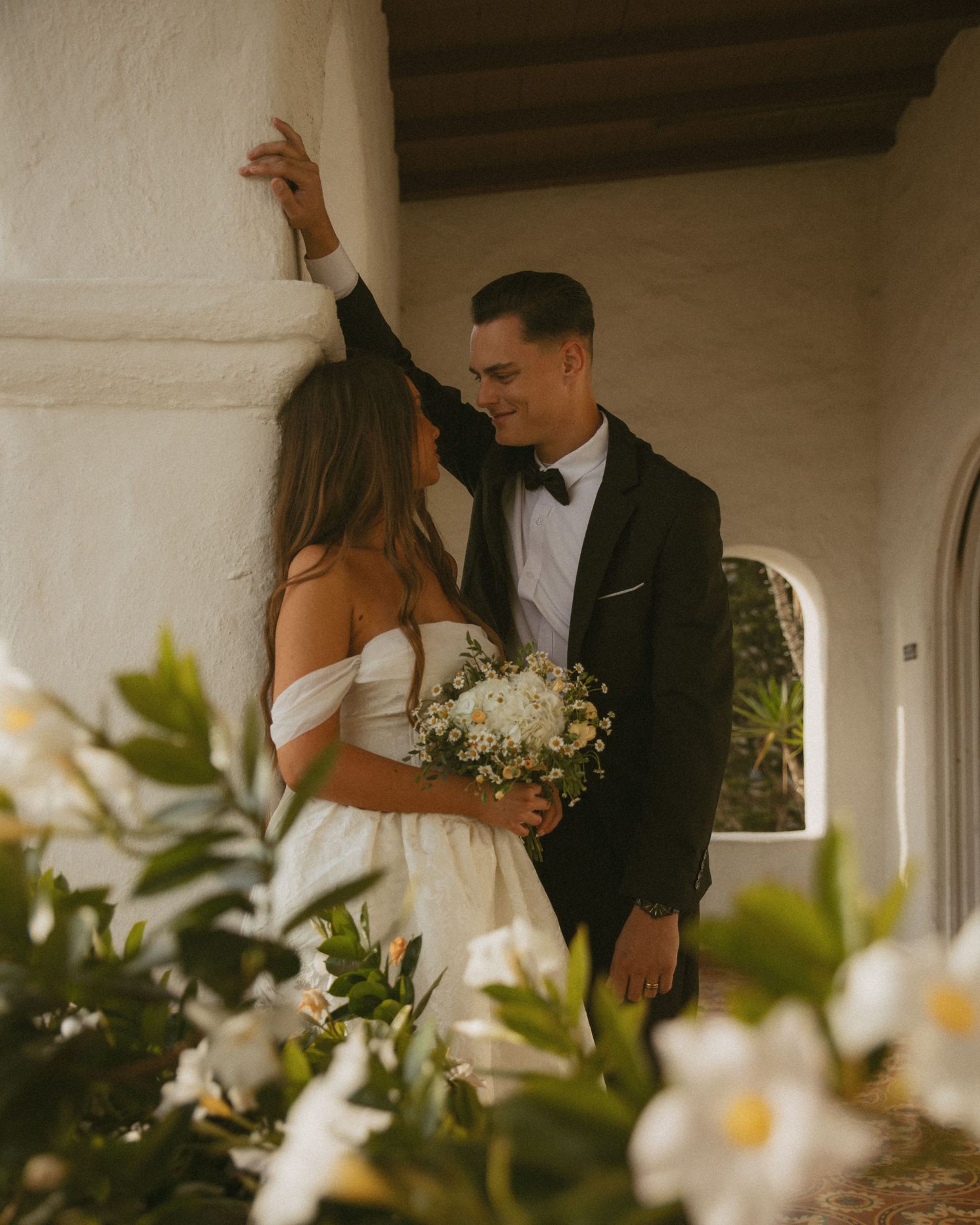 Bride and groom share an intimate moment among white flowers against a whitewashed archway wall.