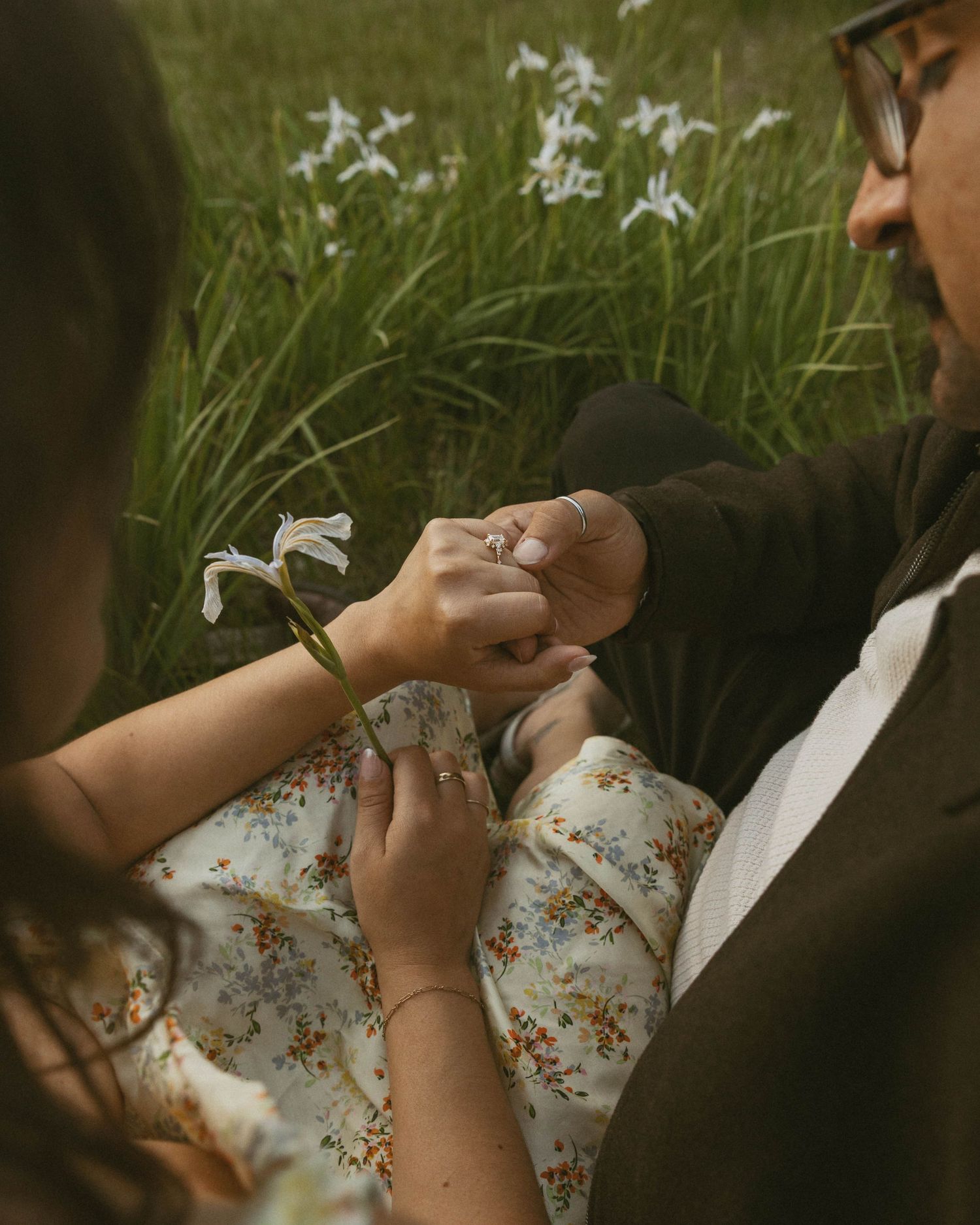 Couple sitting in a wildflower field, holding a white iris bloom together, woman in floral dress wearing a ring.