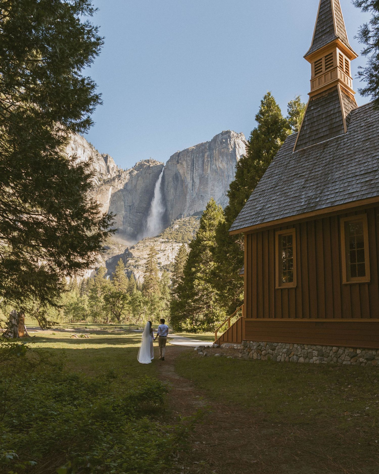 A couple walks toward a historic chapel in Yosemite Valley with a stunning waterfall and granite cliffs in the background.