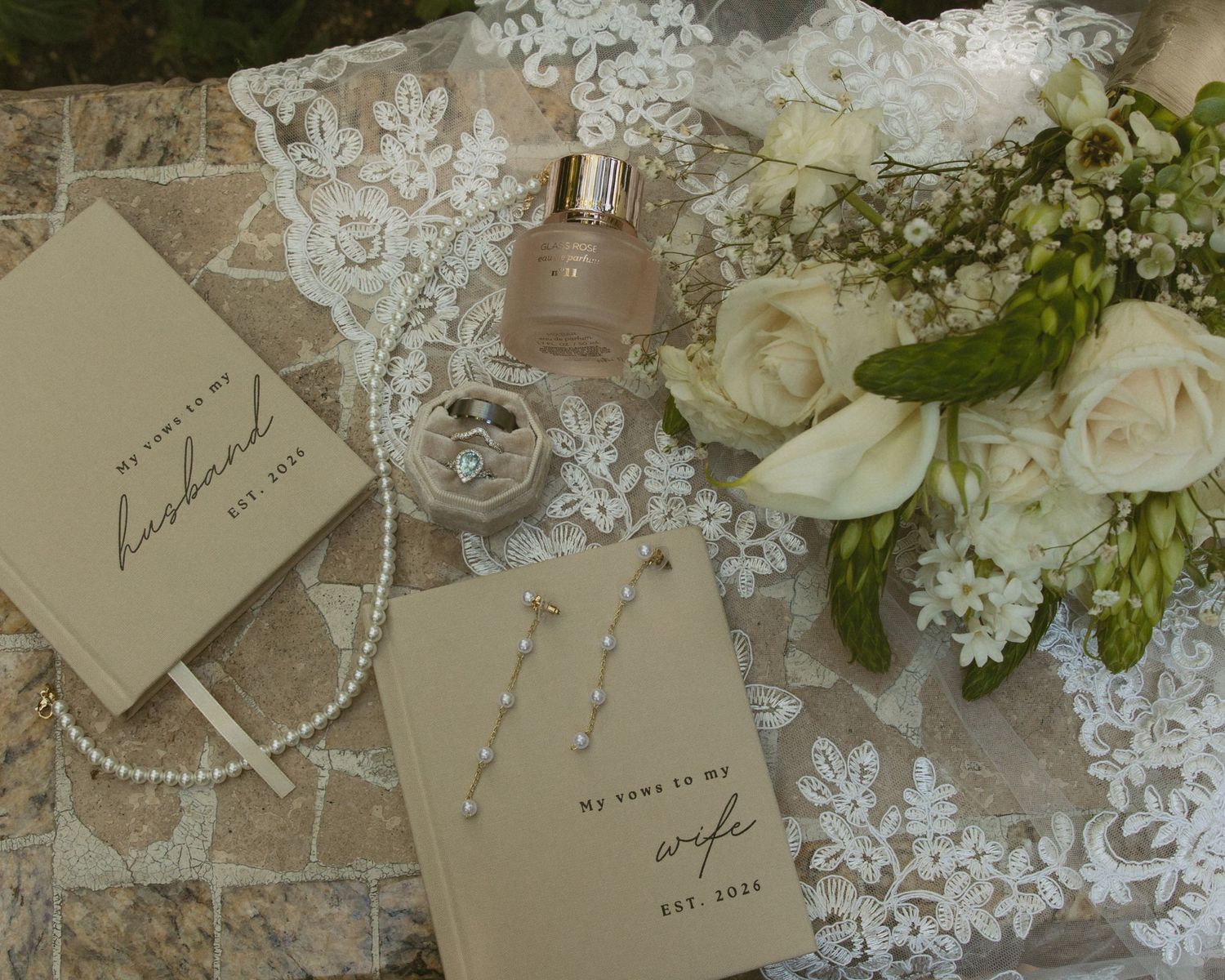 Elegant wedding flat lay with vow books, pearl necklace, earrings, perfume, diamond ring, and white floral bouquet on lace.