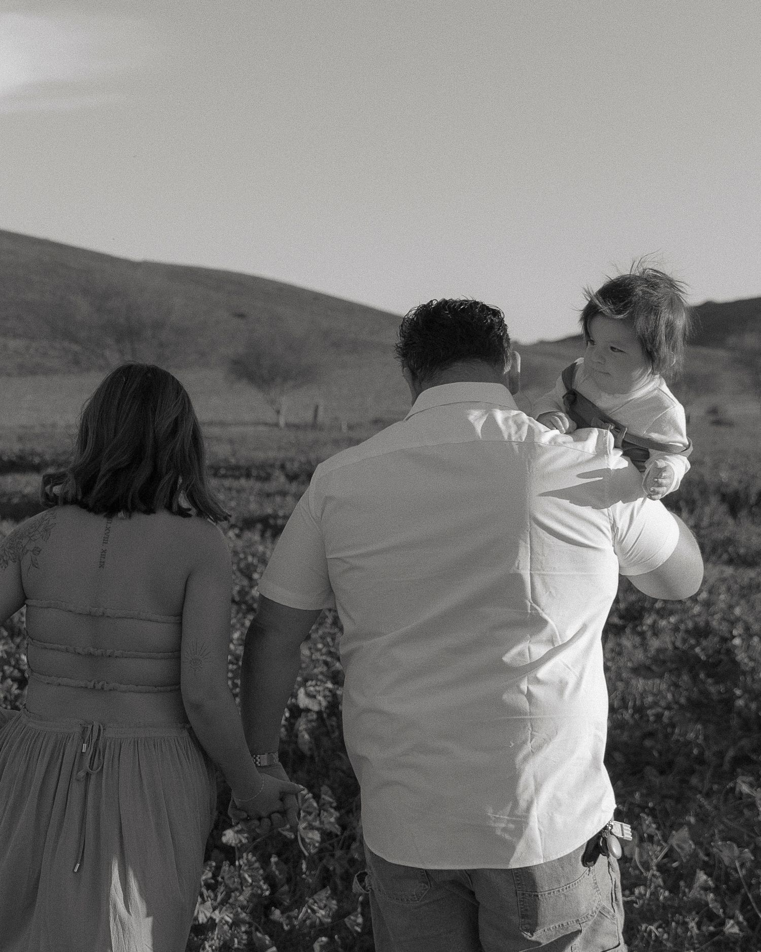 Black and white photo of a family in a vineyard, father holding child while mother walks beside them with hills in background.
