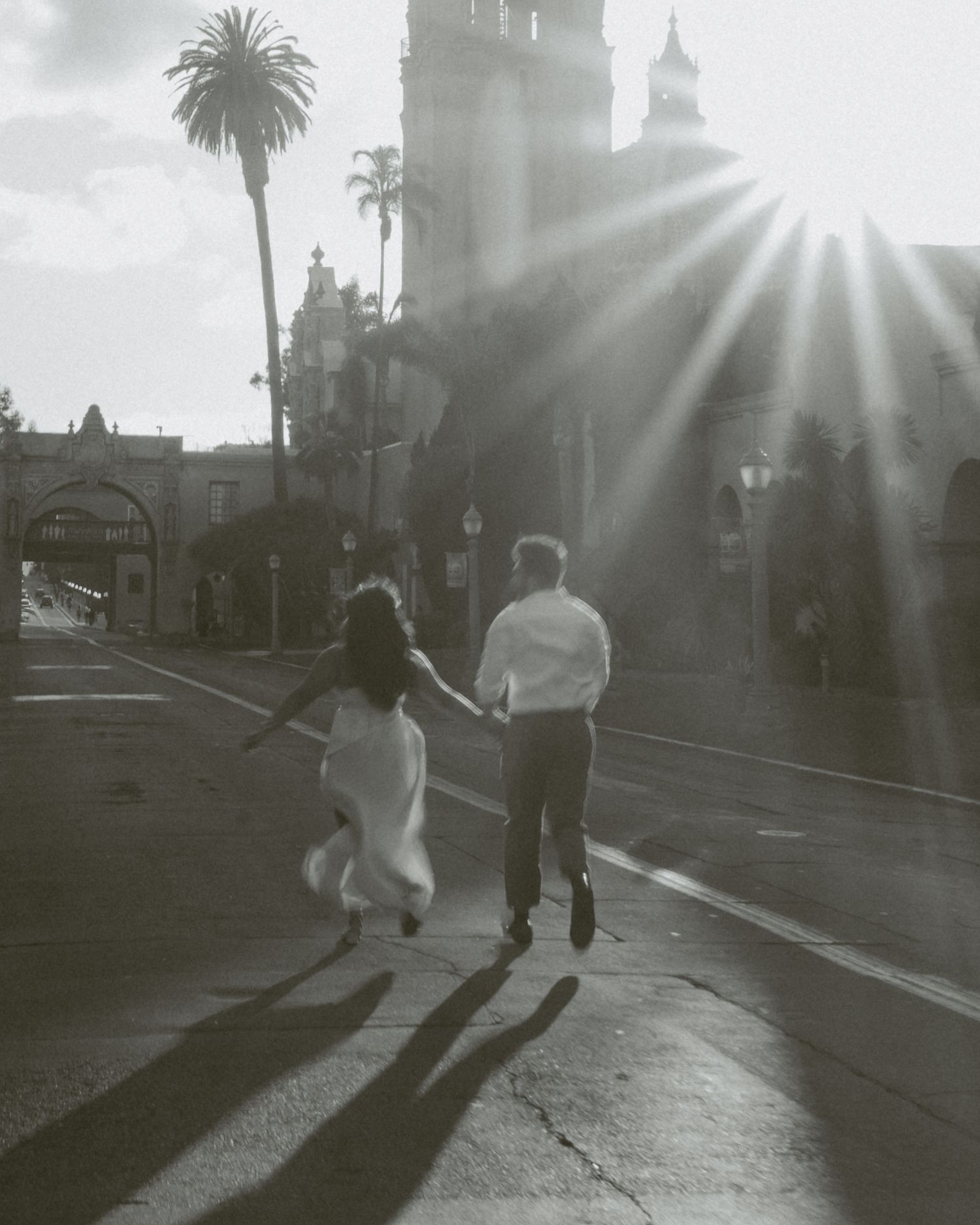 Black and white photo of a couple holding hands running toward sunburst light near a palm tree and historic building.