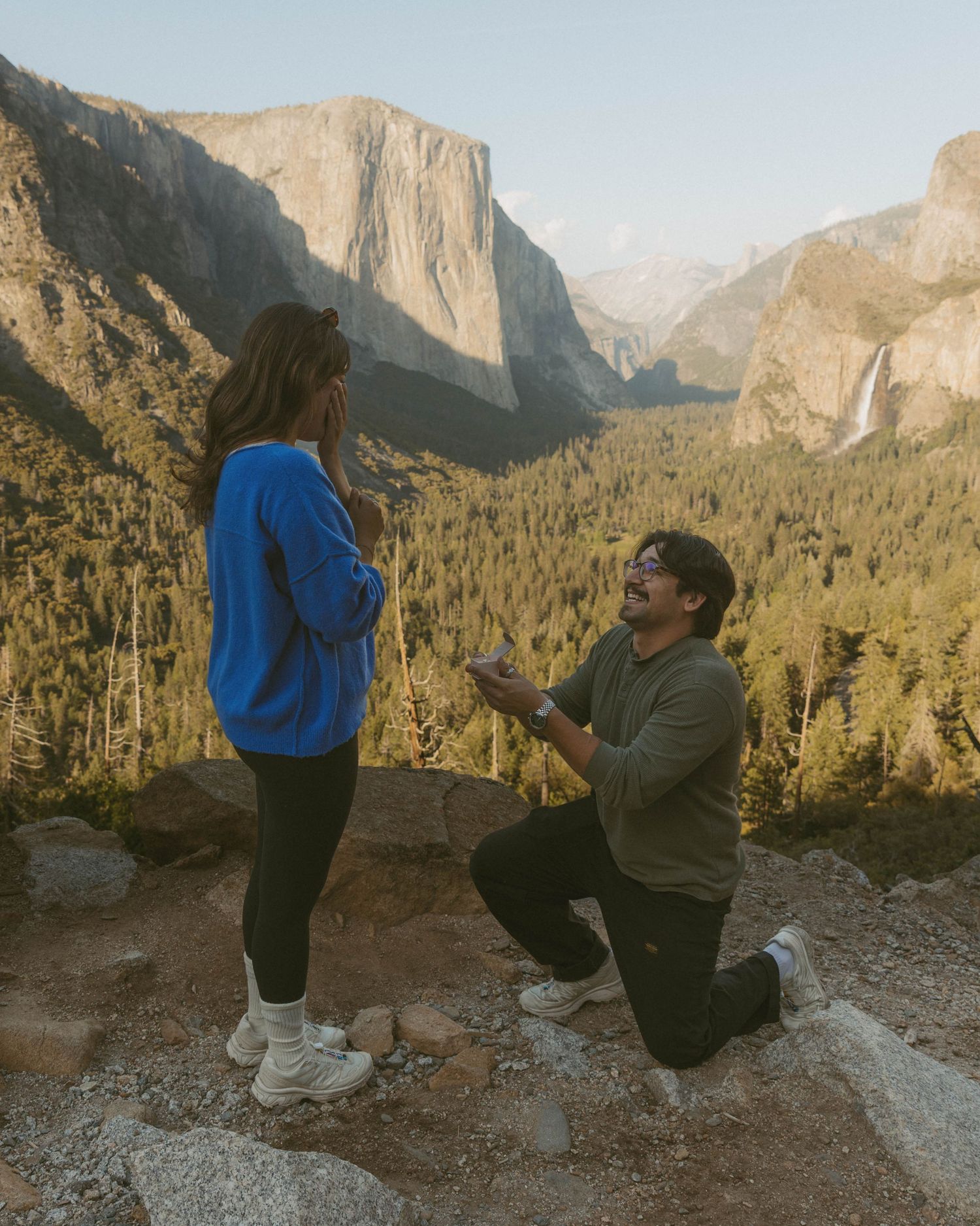 Man proposes on one knee at Yosemite Valley overlook with El Capitan and waterfall backdrop at sunset.