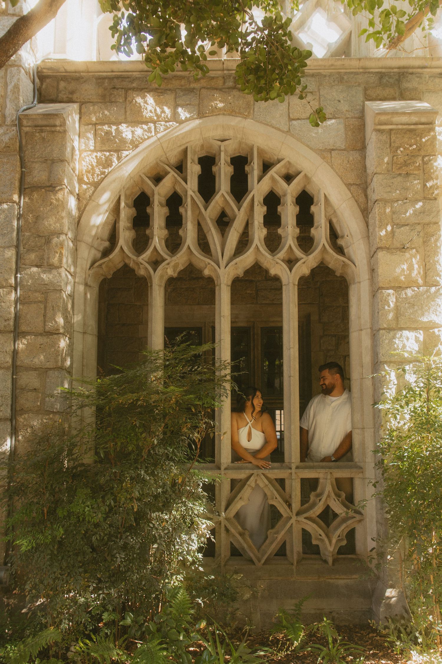 Couple standing inside a Gothic stone archway window surrounded by lush green foliage in warm sunlight.