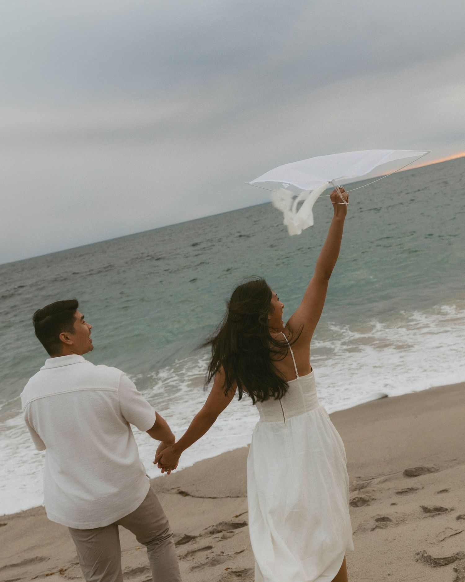 Couple in white outfits holding hands on a beach, woman raising a flowing white fabric toward the cloudy sky.