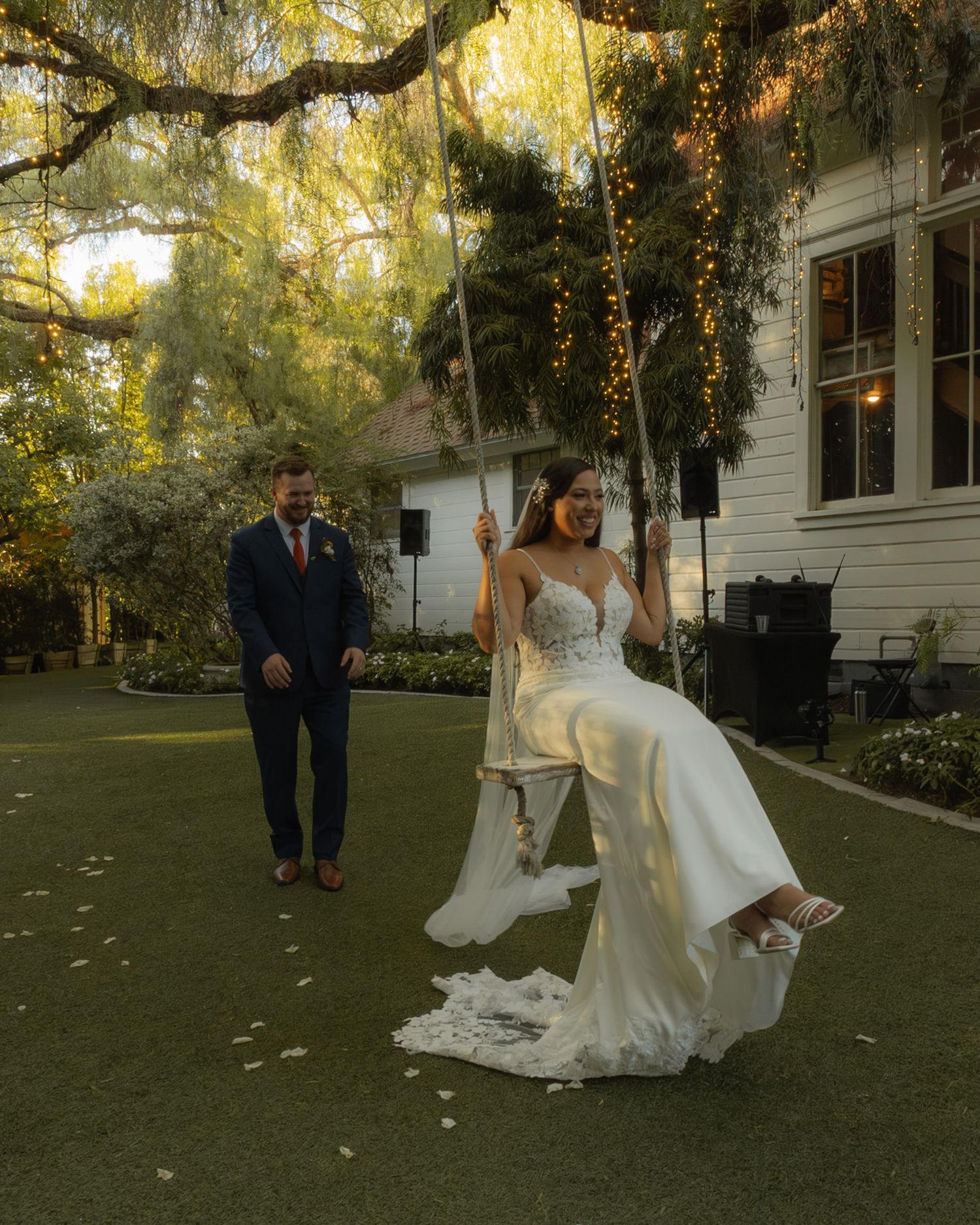 Bride in lace gown sits on garden swing while groom watches at outdoor wedding reception.