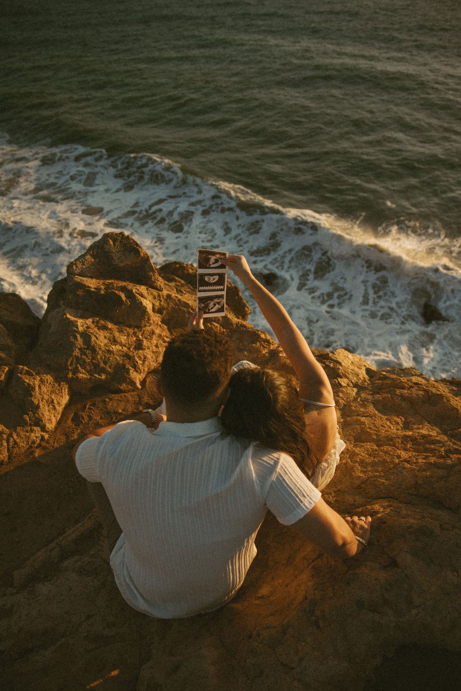 Person in white shirt sits on coastal cliff edge taking photo of ocean waves crashing below during golden hour.