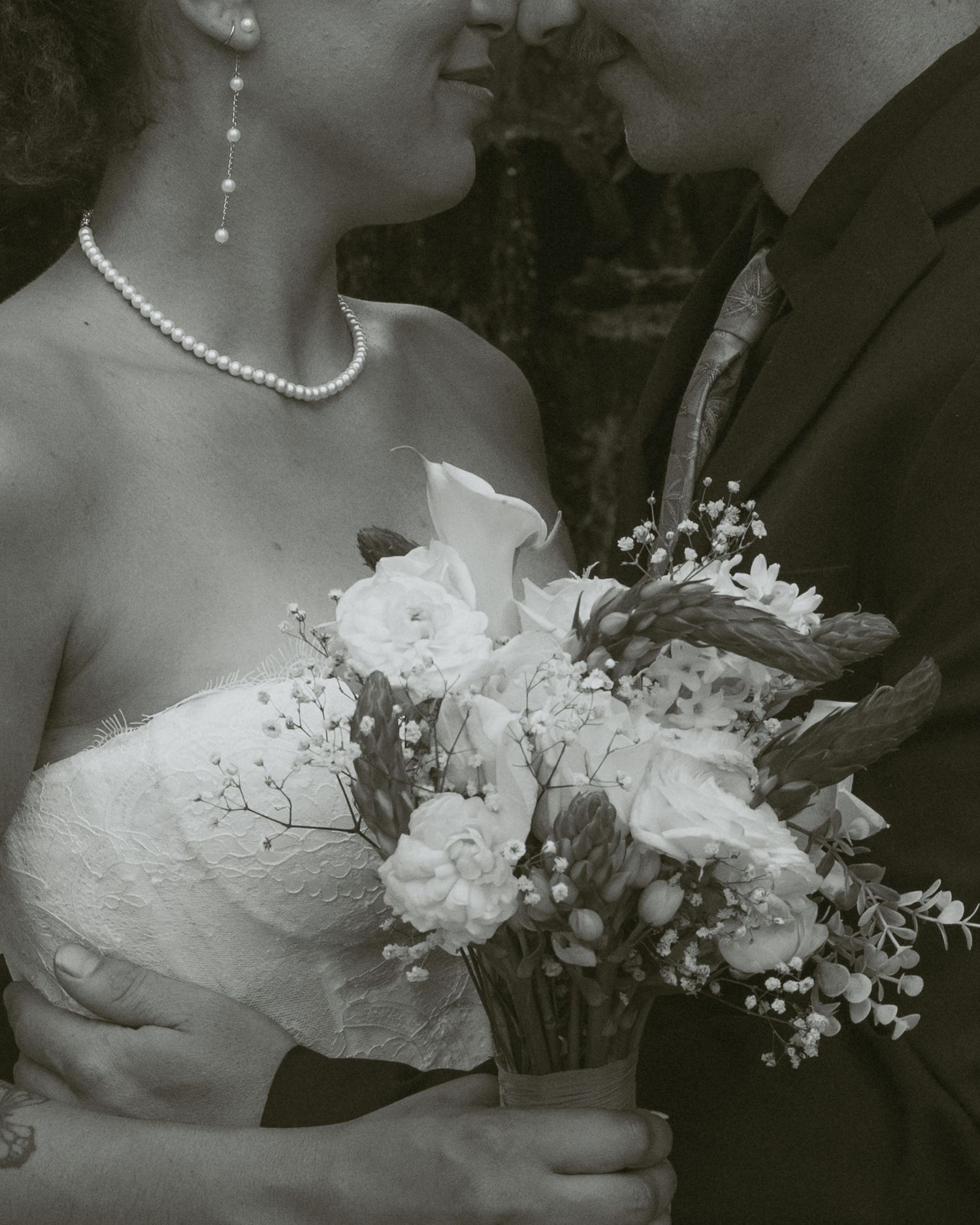 Black and white wedding photo of bride holding floral bouquet with roses, wearing pearl necklace and earrings.