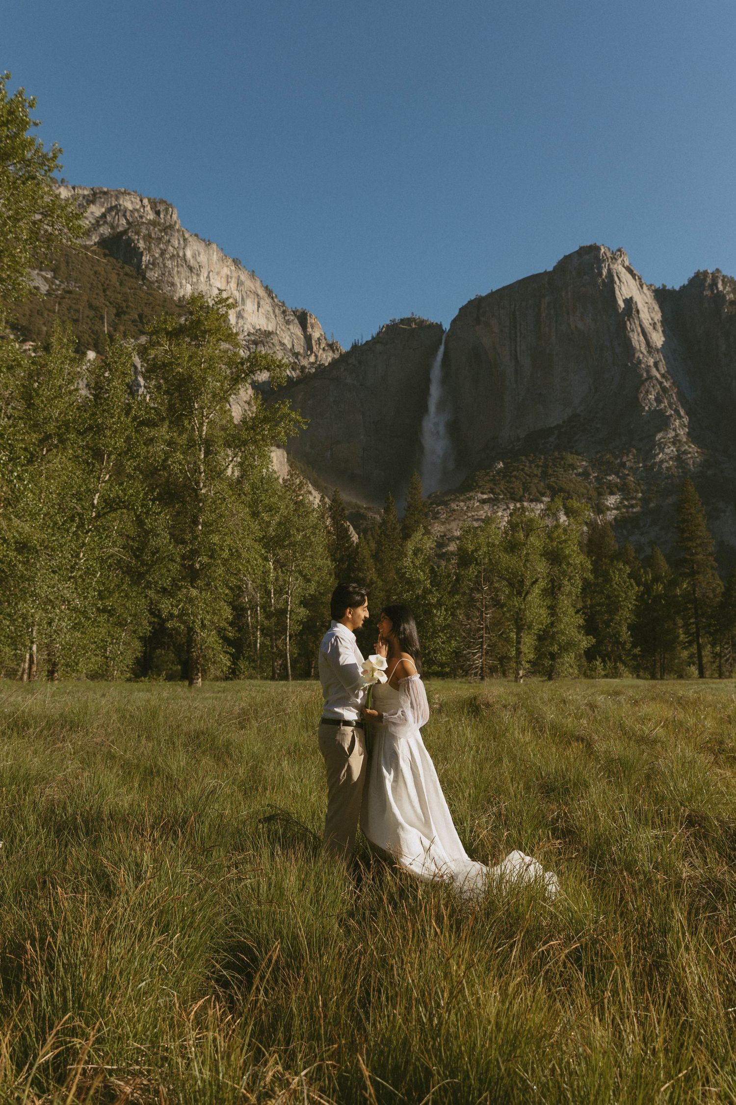 Couple in wedding attire embracing in meadow with Yosemite Falls and granite cliffs in background.