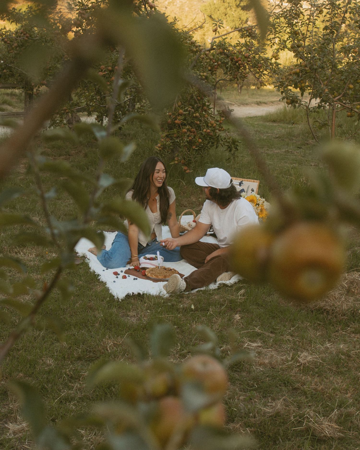 A couple enjoys a romantic picnic on a white blanket surrounded by lush apple trees in a serene orchard setting.