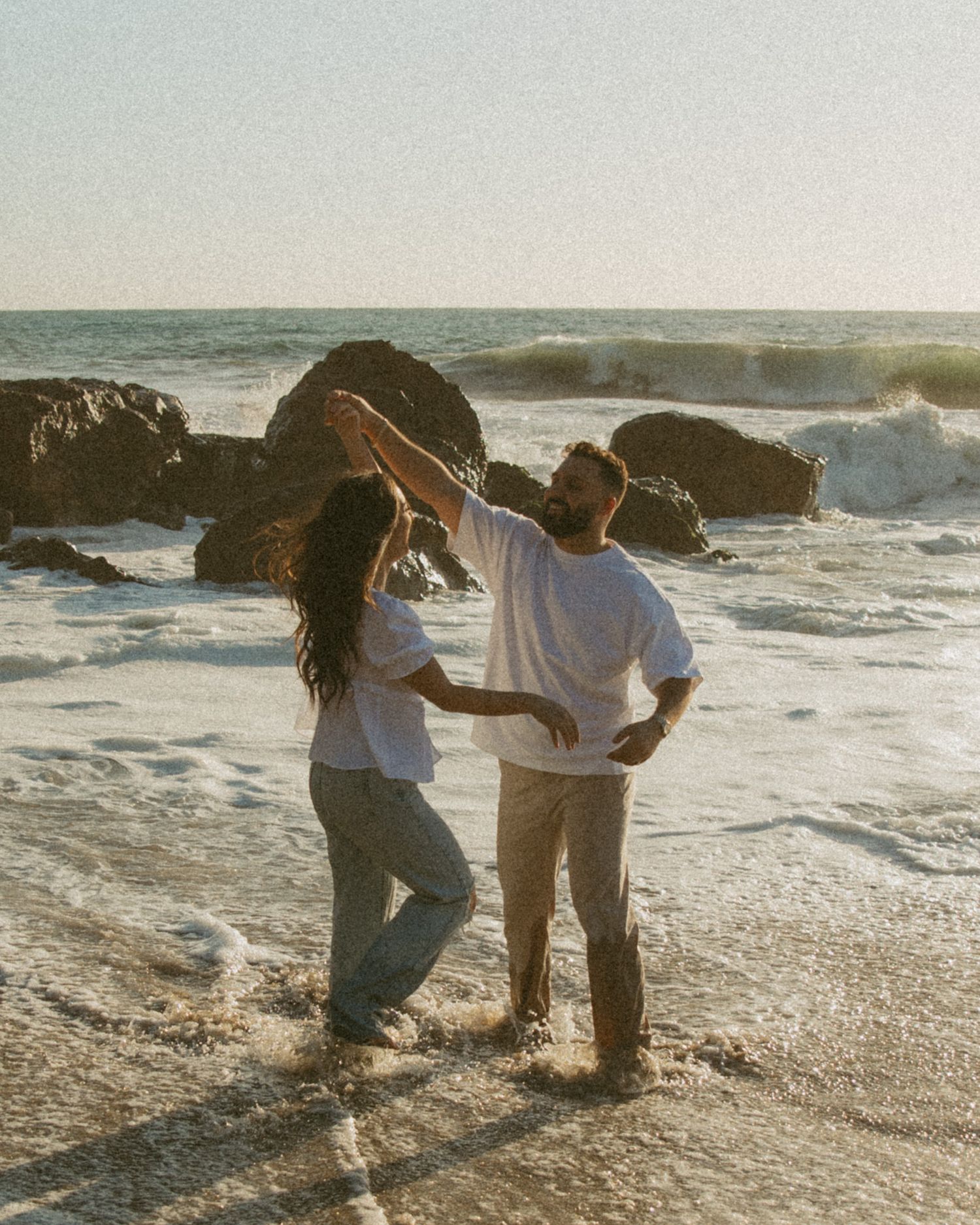 A couple dances together on a rocky beach at sunset with crashing waves and ocean spray in the background.