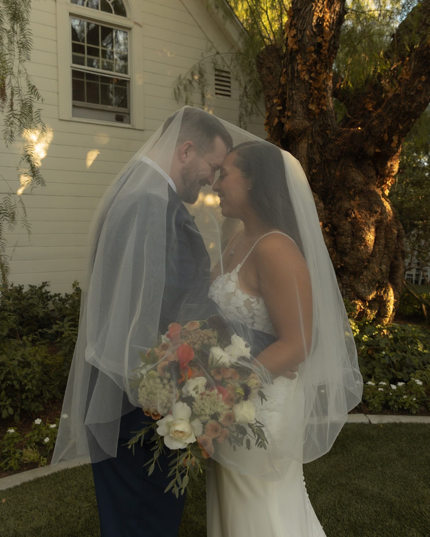 Bride and groom share an intimate moment under a flowing veil, bride holding a floral bouquet outdoors near a white chapel.