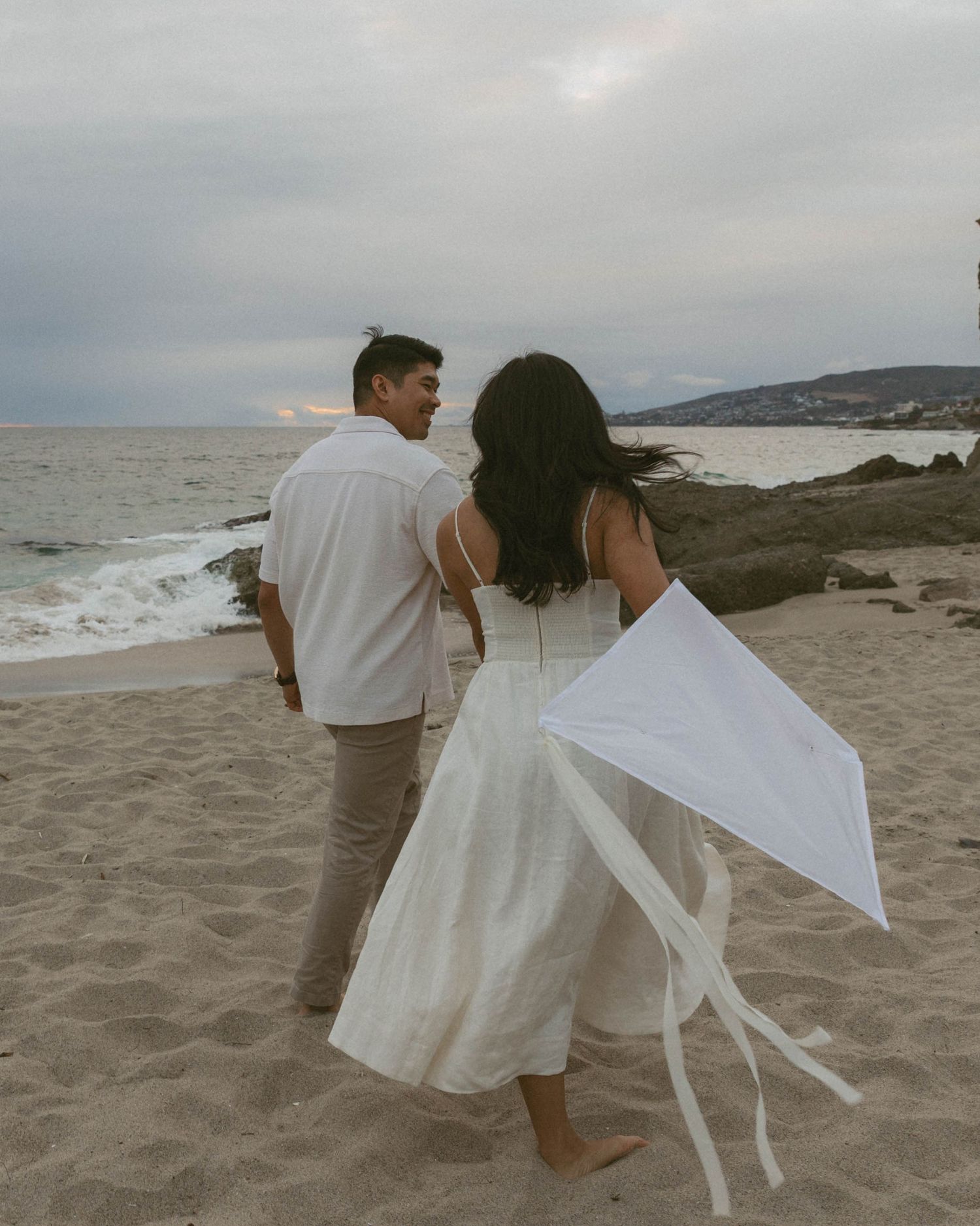Couple in white outfits walking on beach at dusk flying a white kite with ribbons near rocky shoreline.