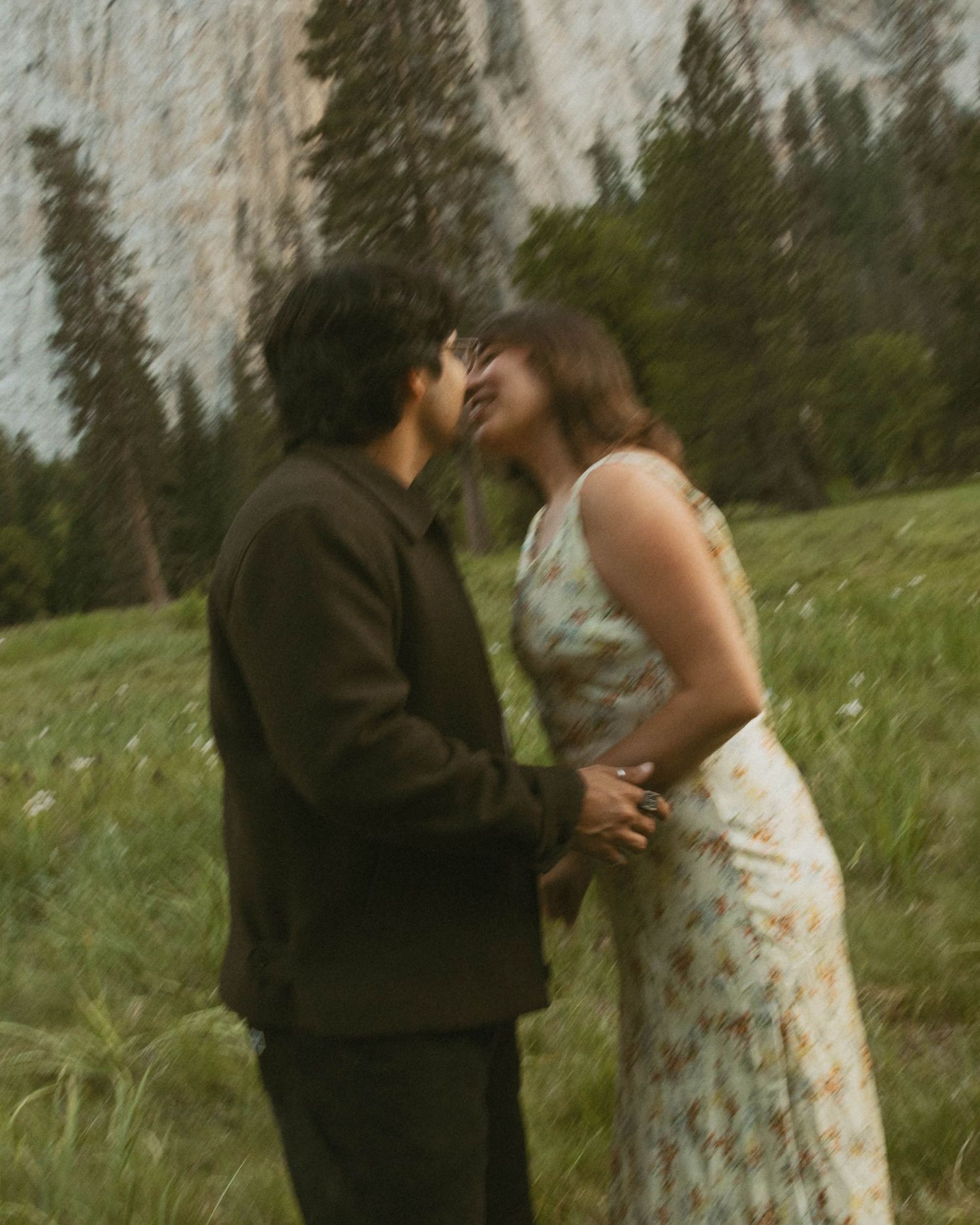 A couple shares a romantic almost-kiss moment in a misty meadow with pine trees and a mountain cliff backdrop.