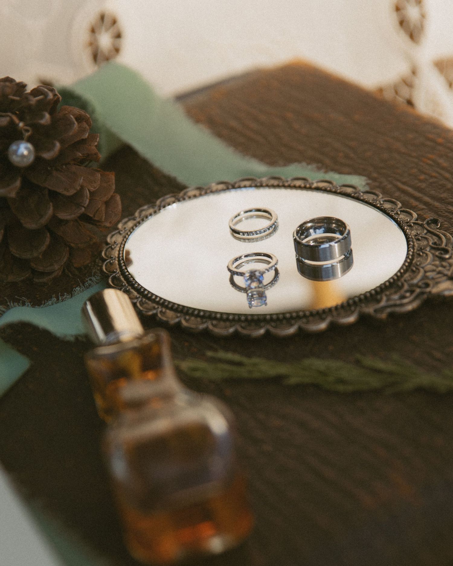 Elegant wedding rings displayed on ornate white tray with pine cone, green ribbon, and amber perfume bottle on dark wood surface.