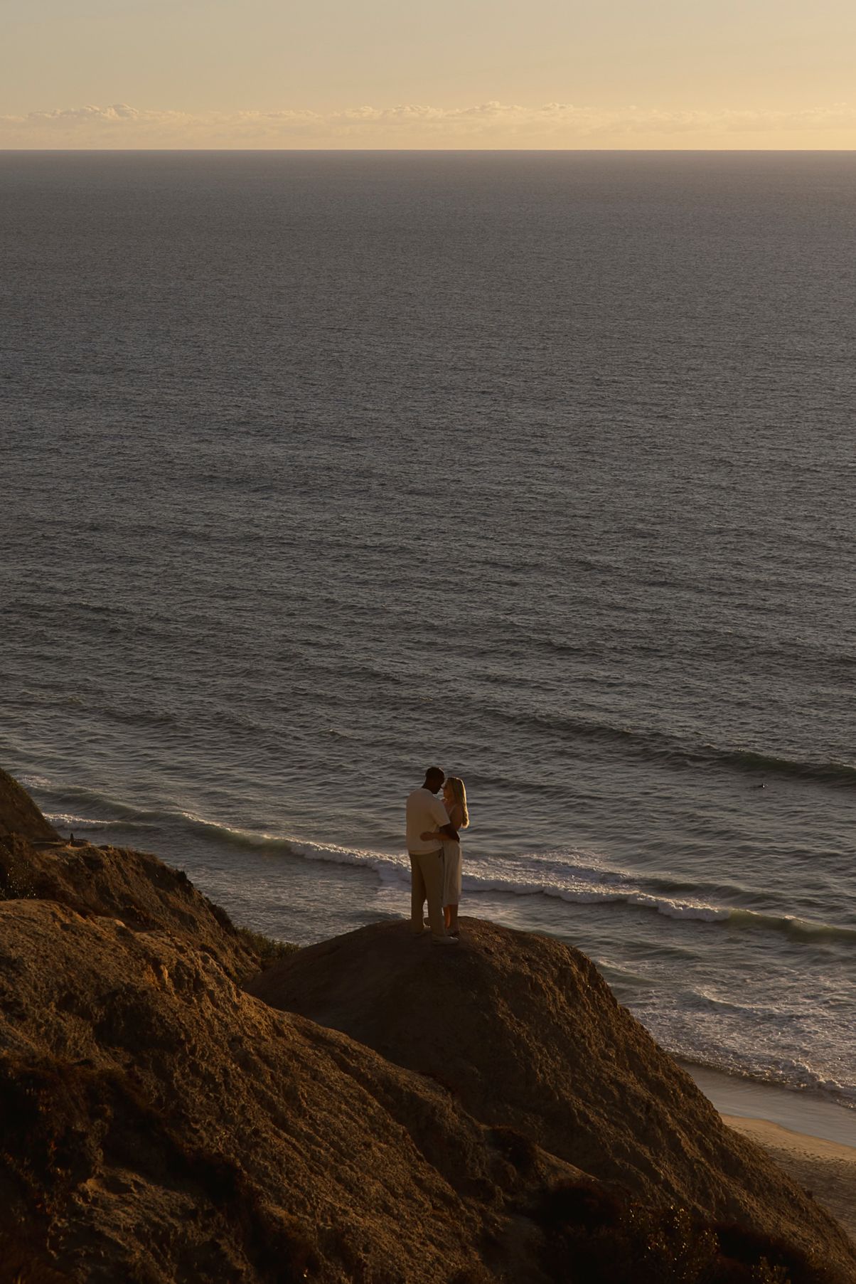 Two silhouettes stand atop a coastal cliff overlooking the ocean during a golden sunset.