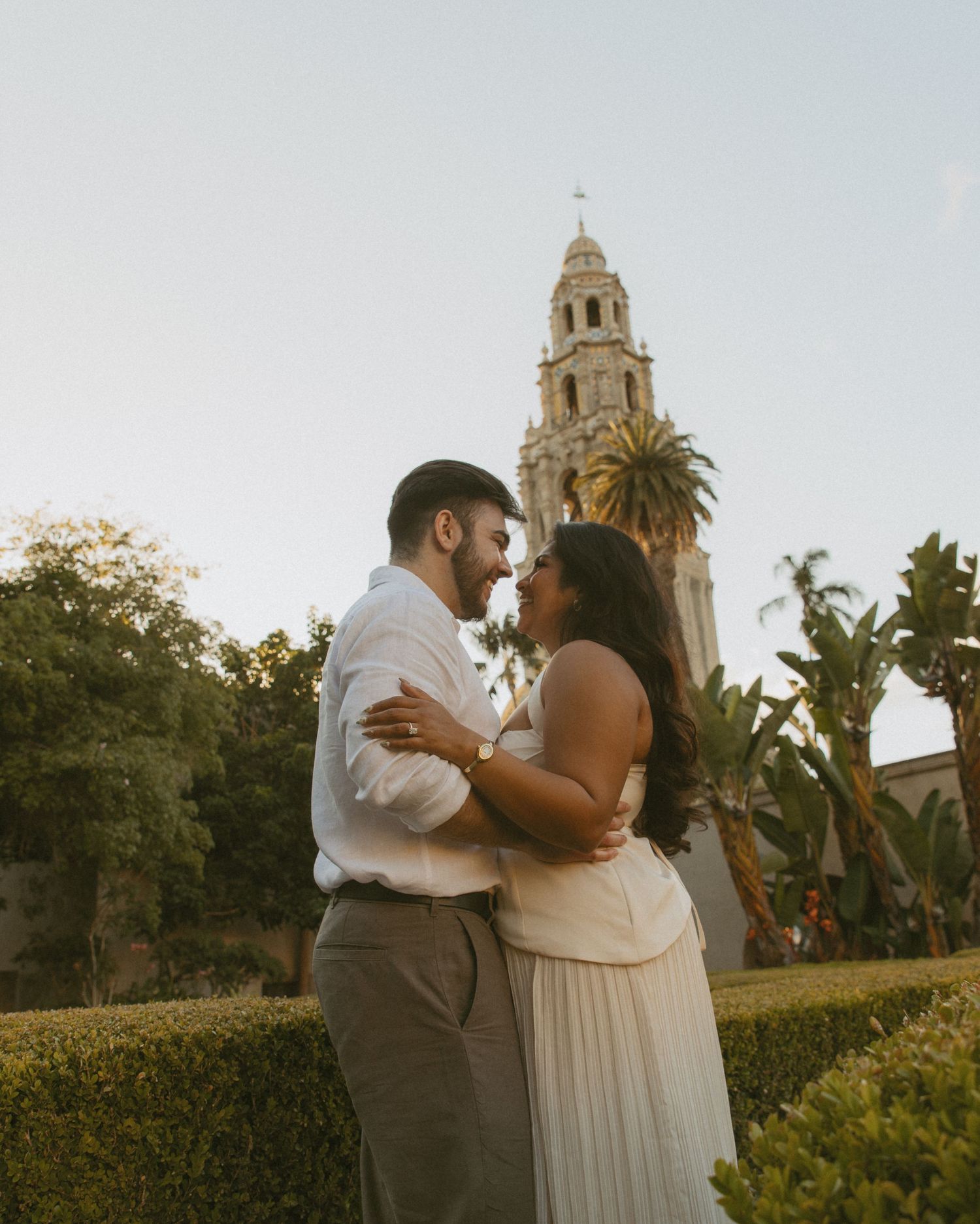 Couple embracing and smiling at each other in Balboa Park with a historic tower and palm trees in the background.