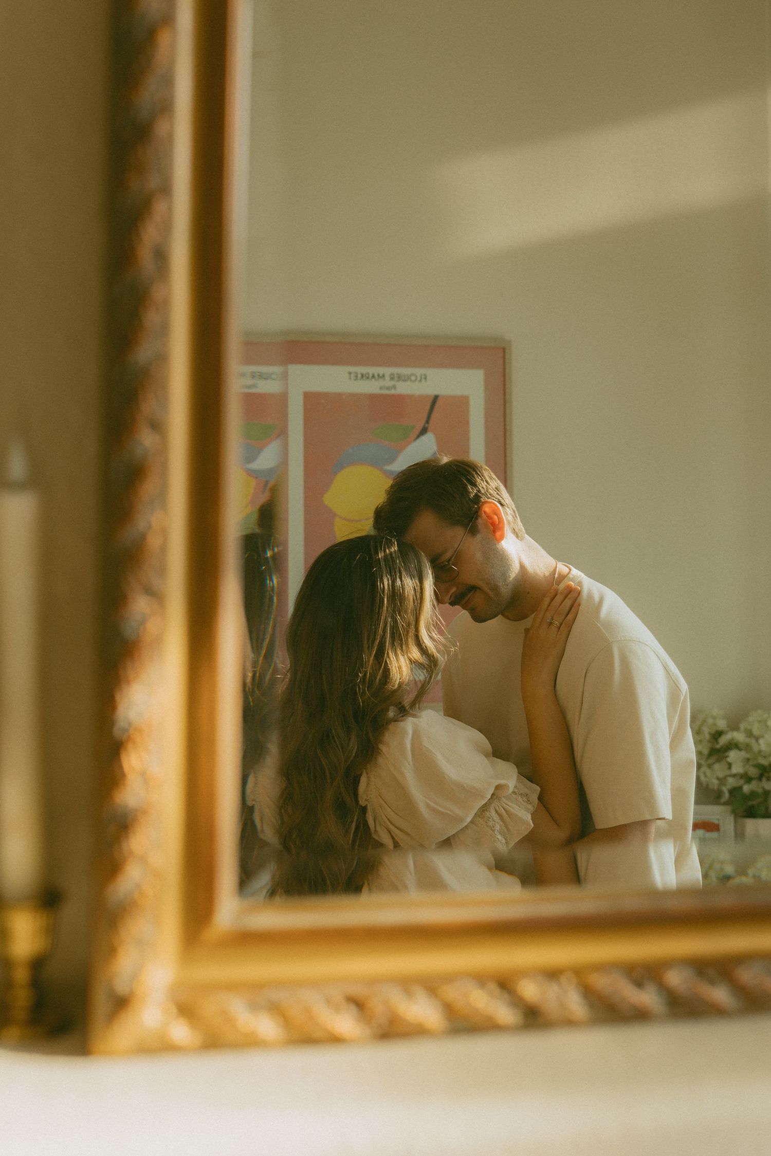 Couple sharing a romantic embrace reflected in a gold ornate mirror with warm sunlight in a cozy room.