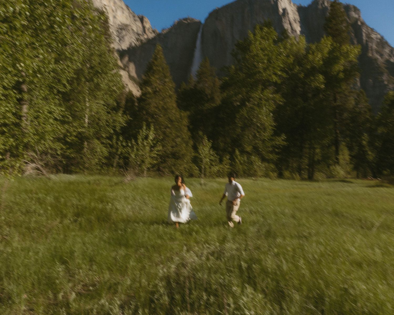 Couple in white attire running through a green meadow with tall trees and a waterfall in Yosemite Valley.