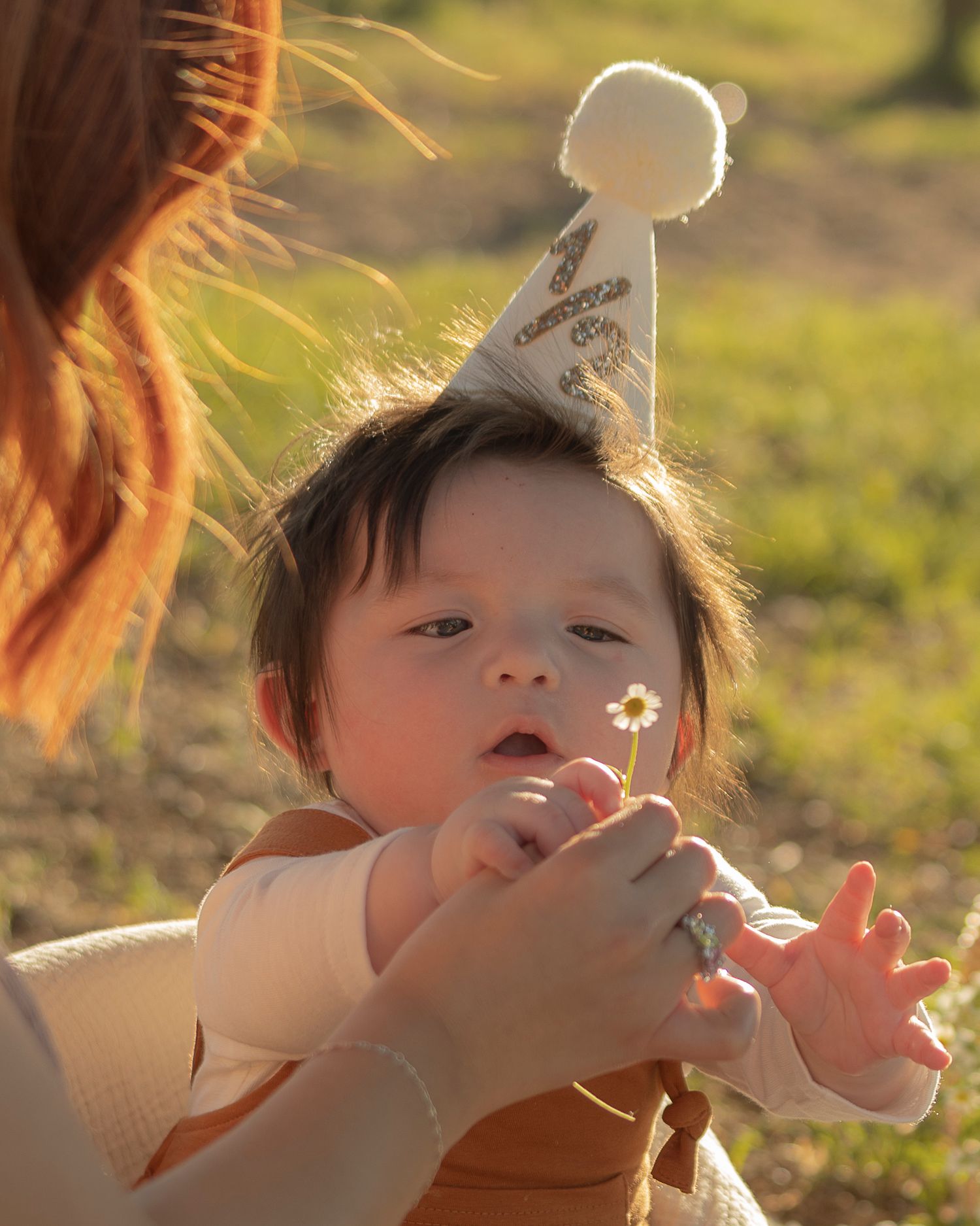 A baby wearing a birthday party hat holds a small flower while being held by a woman outdoors in golden sunlight.