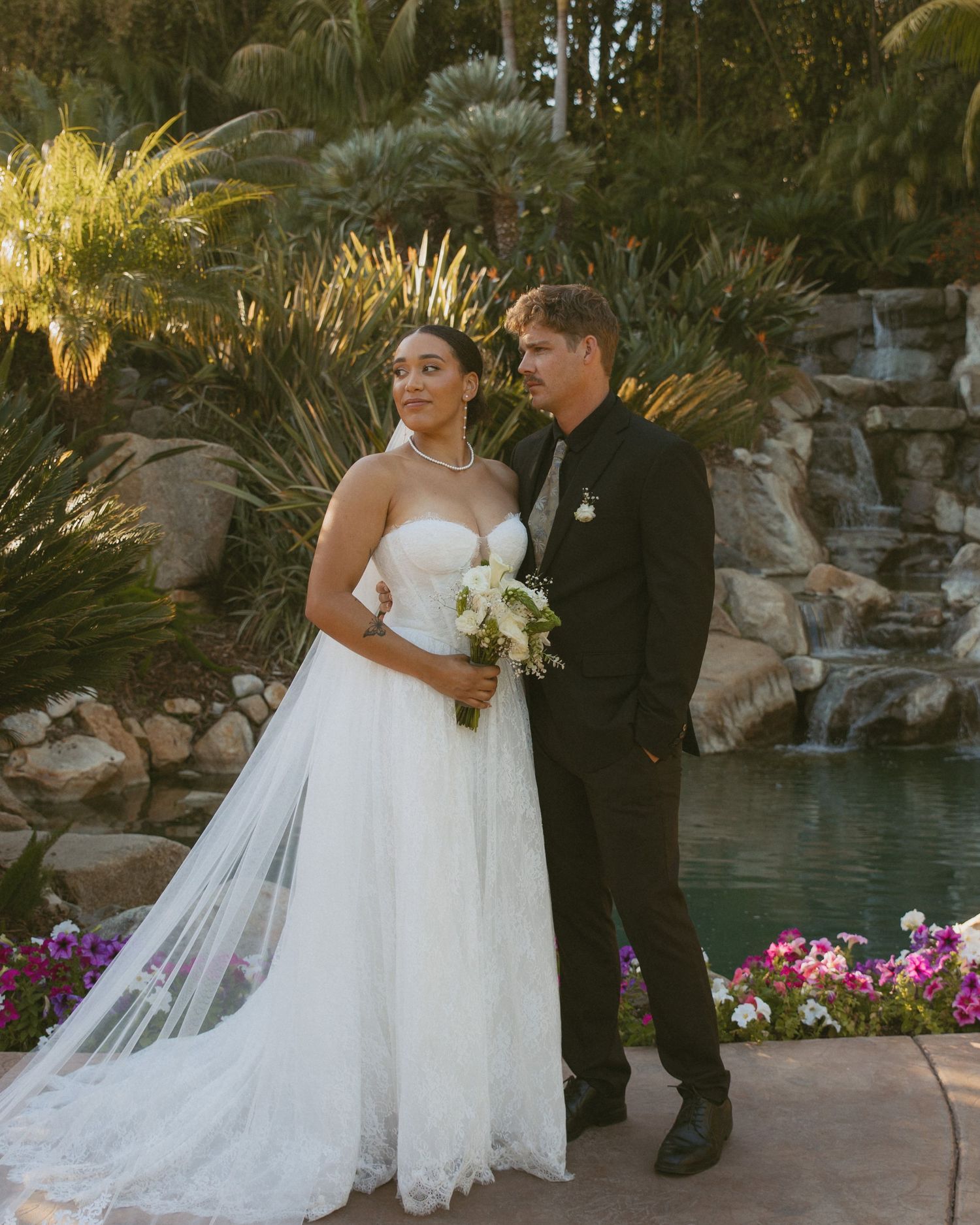 Bride and groom pose together by waterfall garden backdrop at golden hour wedding reception.