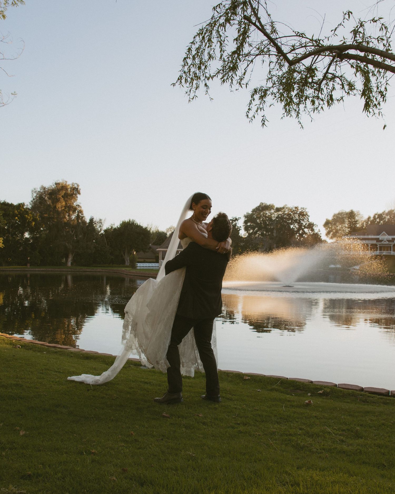Groom lifting bride in white wedding dress by a serene lake with fountain at golden hour.