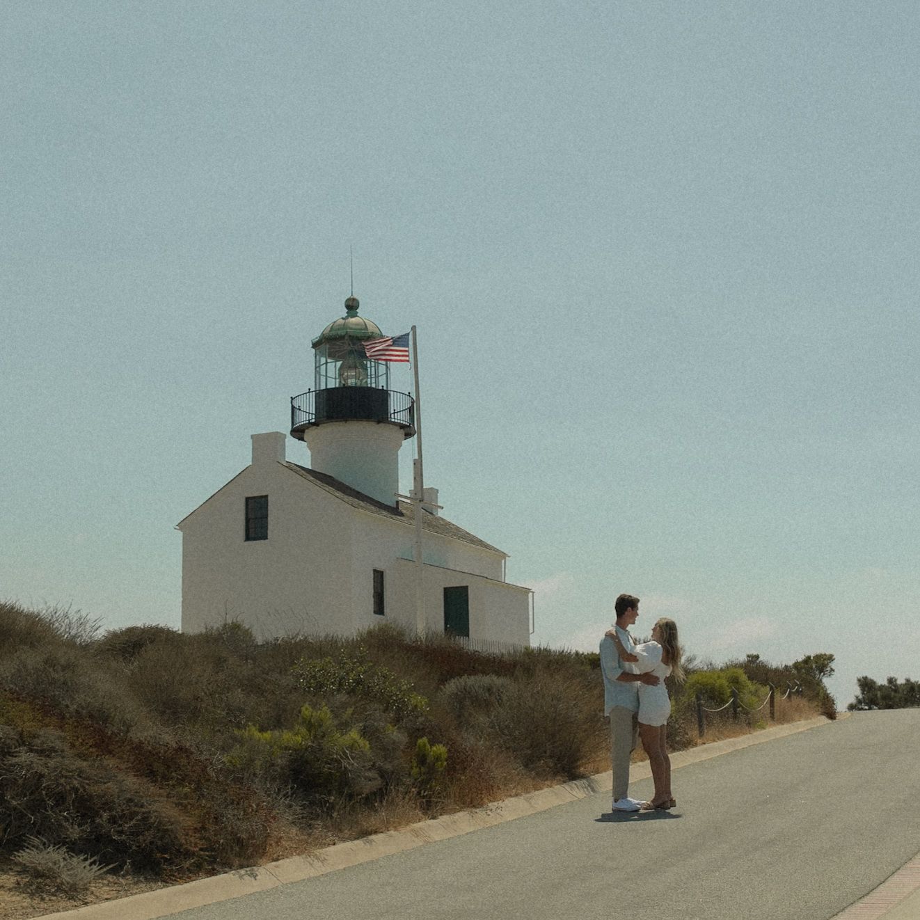 Couple embracing near a white lighthouse with an American flag on a sunny coastal road.