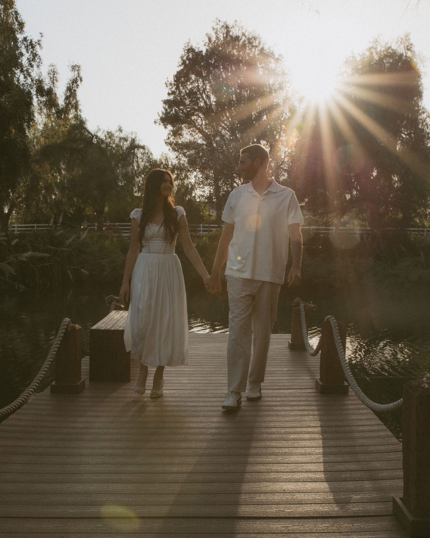 Couple holding hands on a wooden dock at golden hour, dressed in white, with sunburst and lush trees in the background.