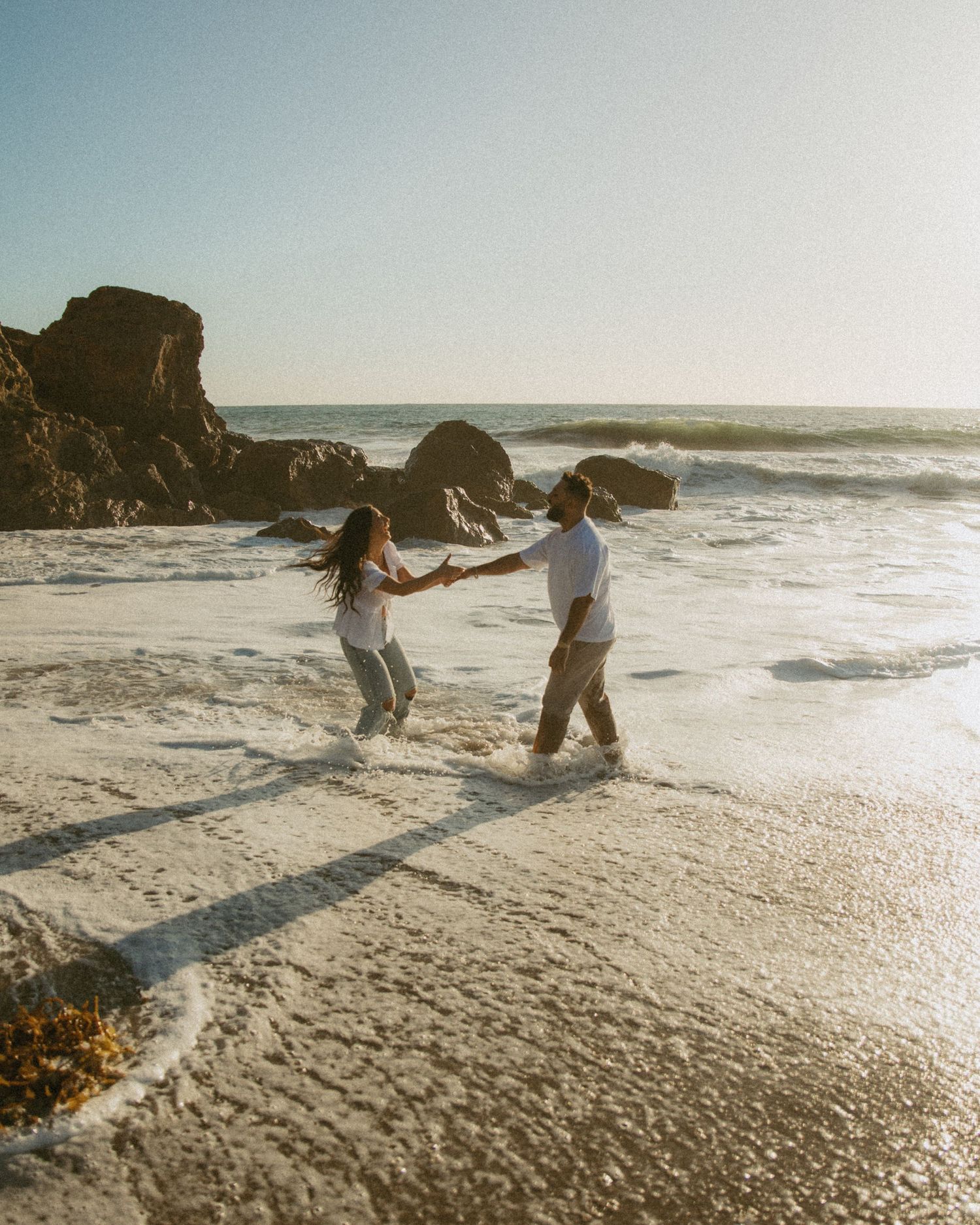 Two people holding hands and spinning together on a sunny beach with ocean waves and rocky cliffs in the background.