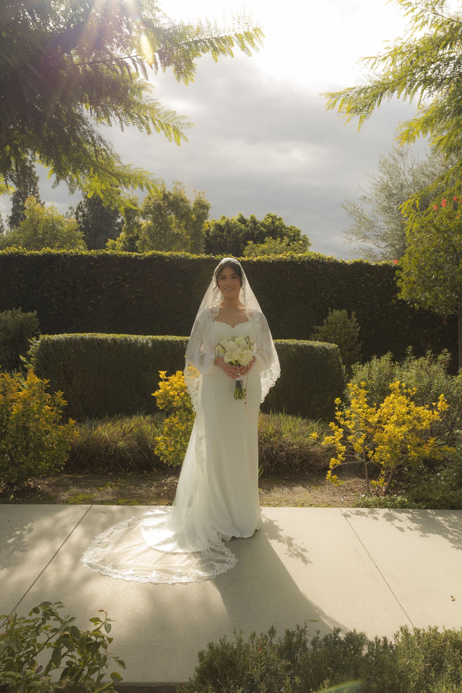 Elegant bride in white gown and veil holding bouquet in sunlit formal garden hedge setting.