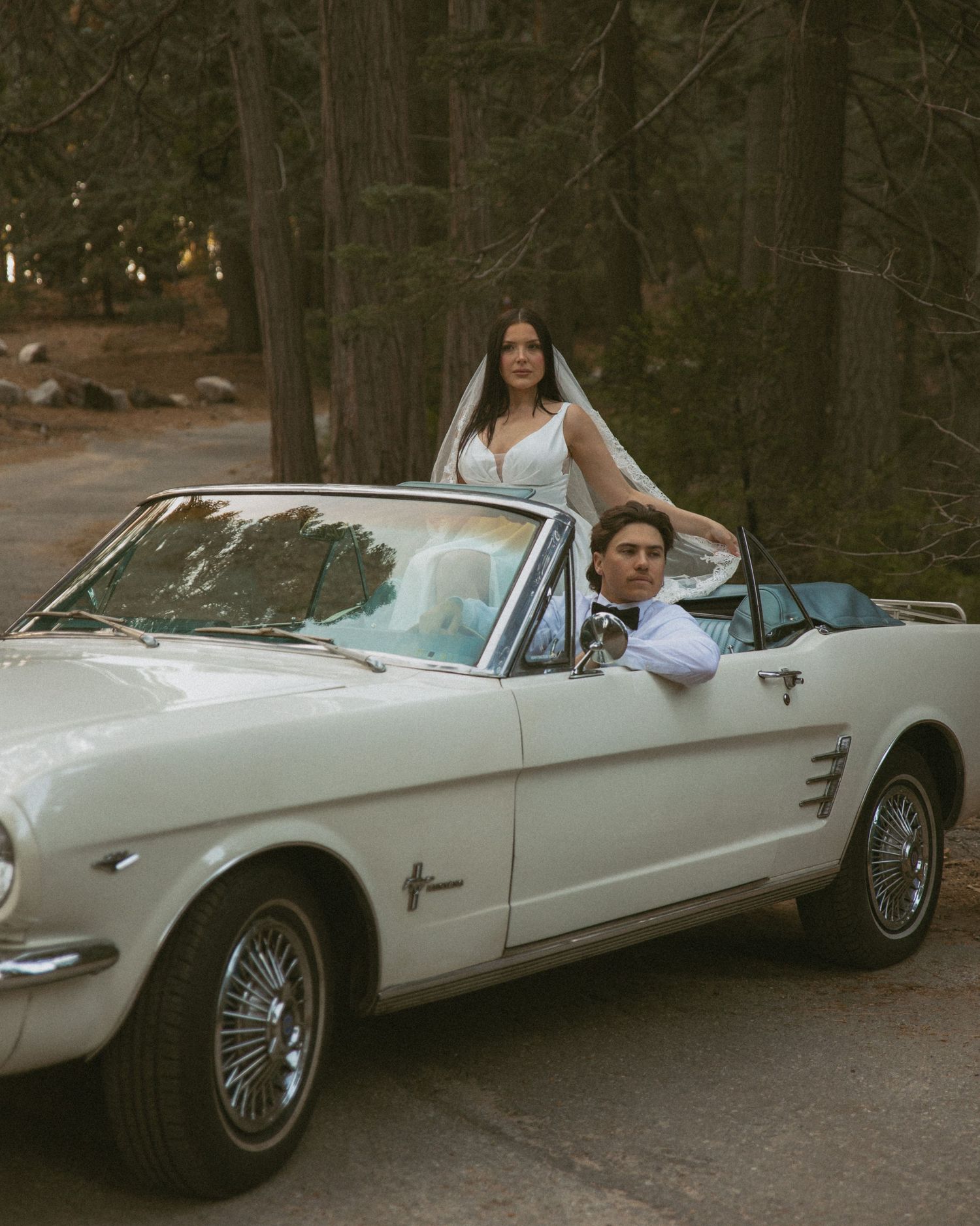 Bride in white veil stands behind groom seated in classic white Ford Mustang convertible on a forest road.