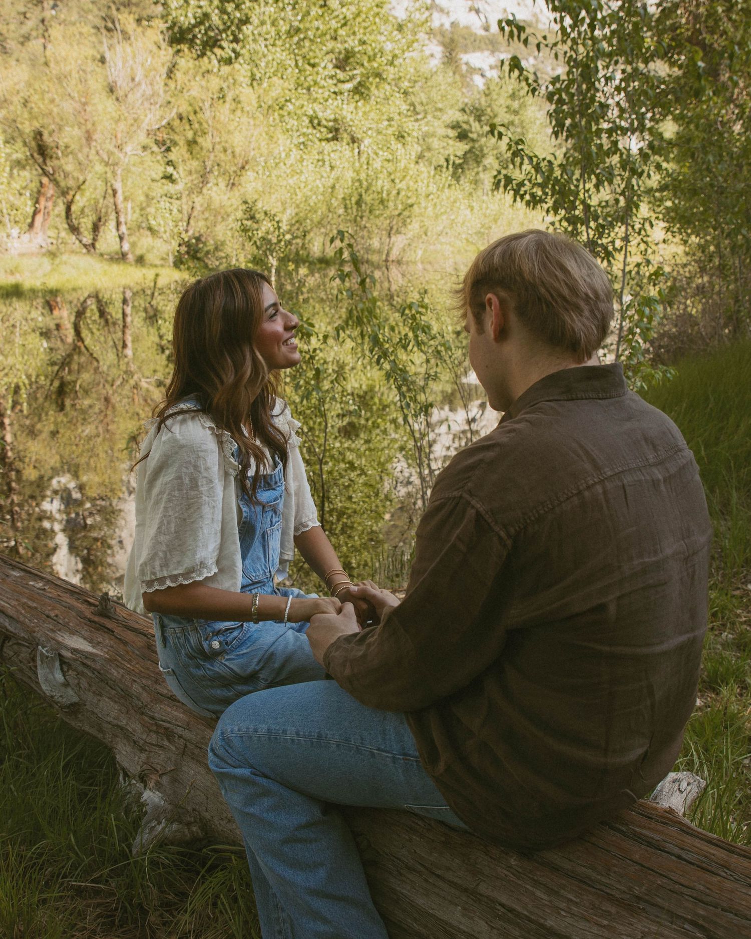 Couple sitting on a fallen log by a reflective pond, surrounded by lush green trees, holding hands and smiling.