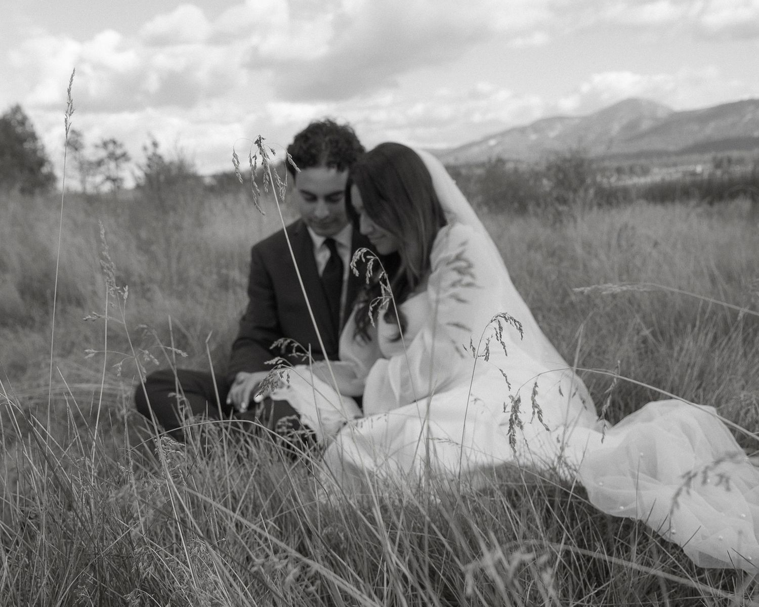 A couple in wedding attire sits together in a grassy field with mountains in the background.