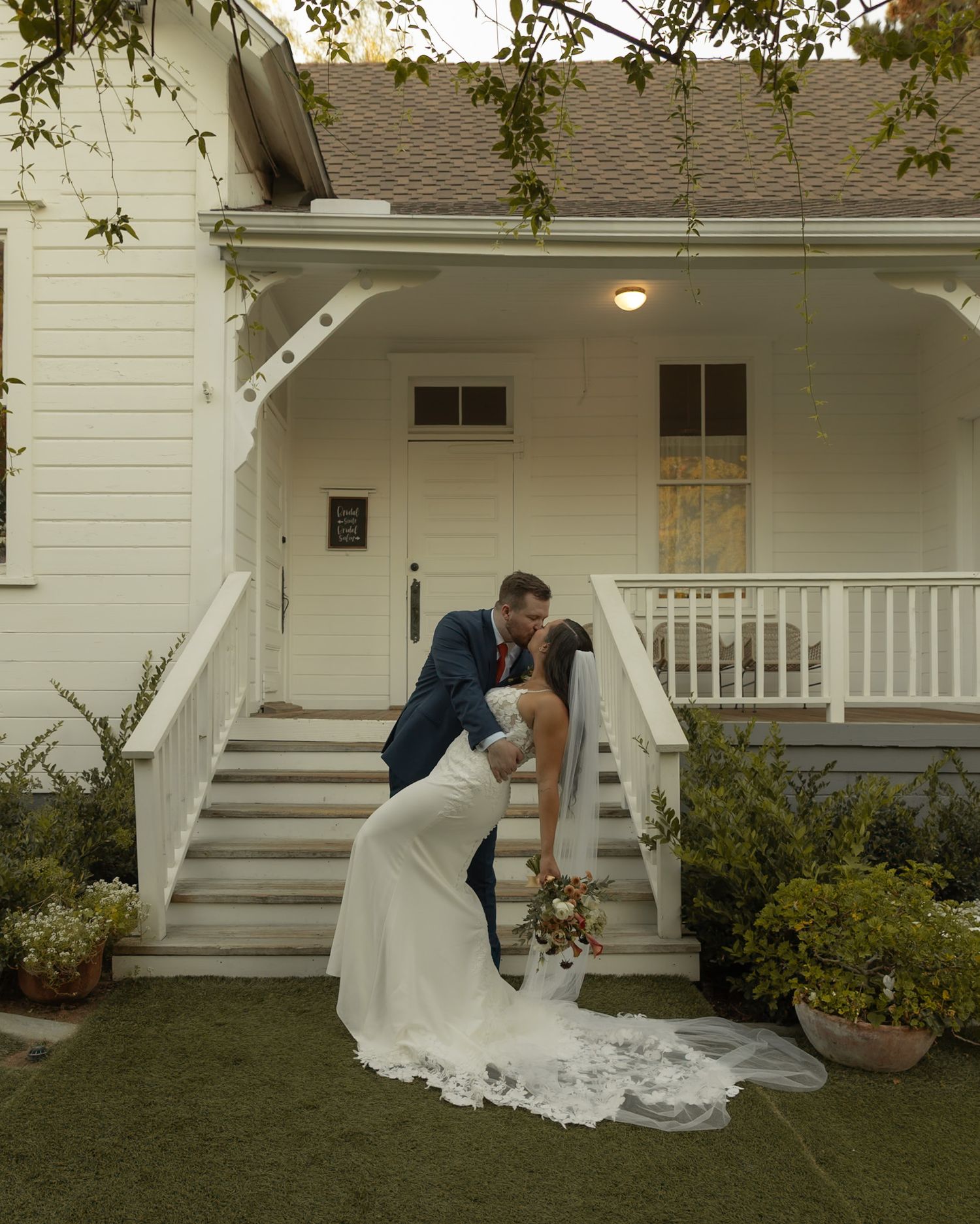 Bride and groom share a romantic kiss on the steps of a charming white farmhouse wedding venue.