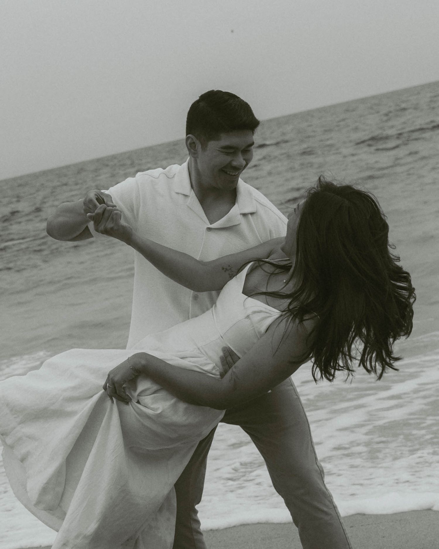 Black and white photo of a couple dancing and laughing together on a beach with ocean waves behind them.