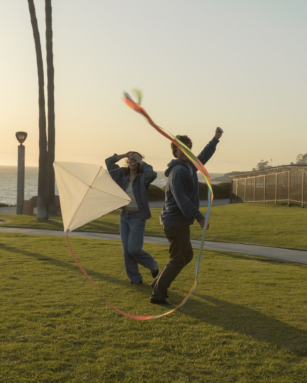 Two people flying a colorful kite with long ribbons on a grassy park near the ocean at sunset.