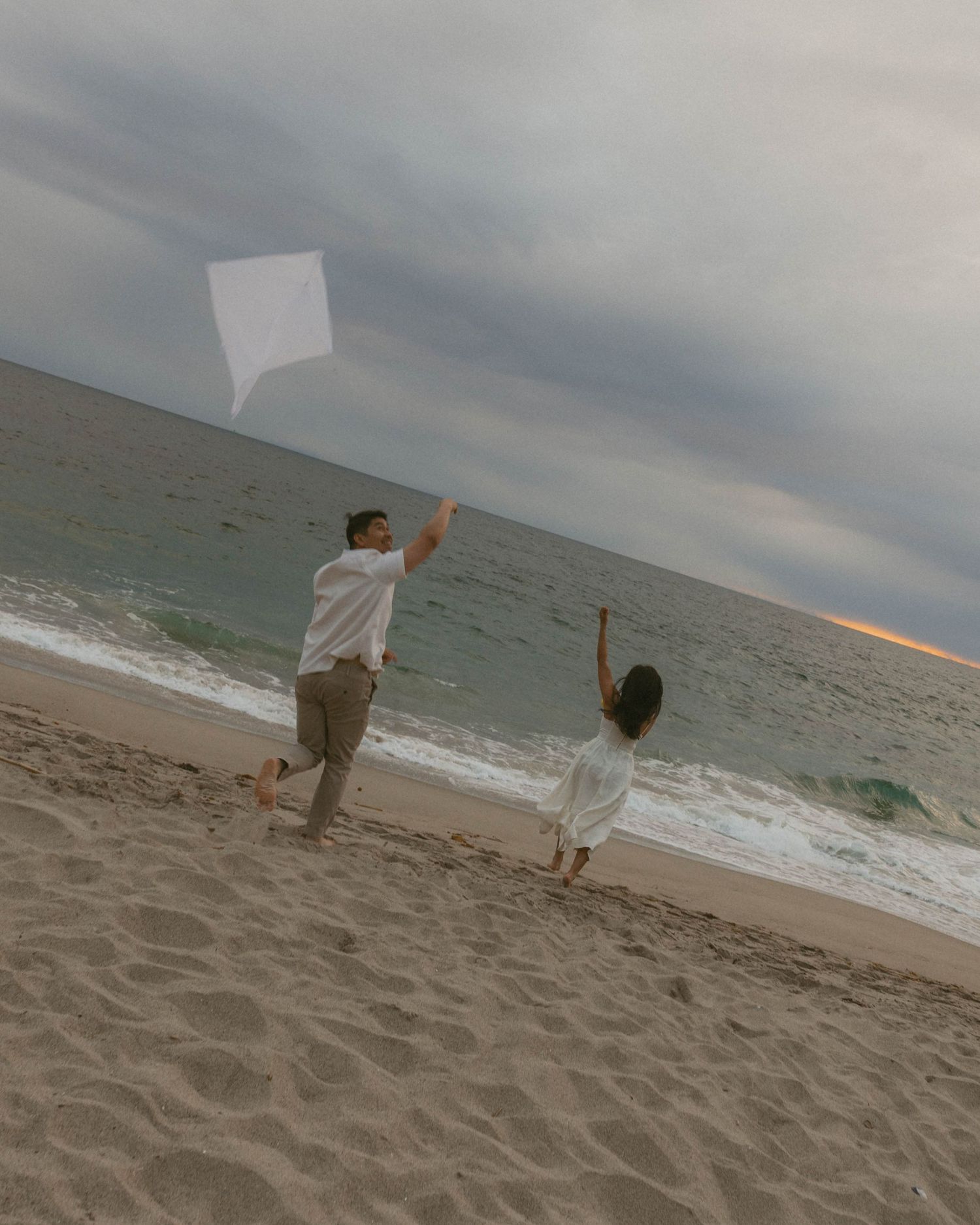 A man and child fly a white kite together on a sandy beach at dusk with cloudy skies and ocean waves.