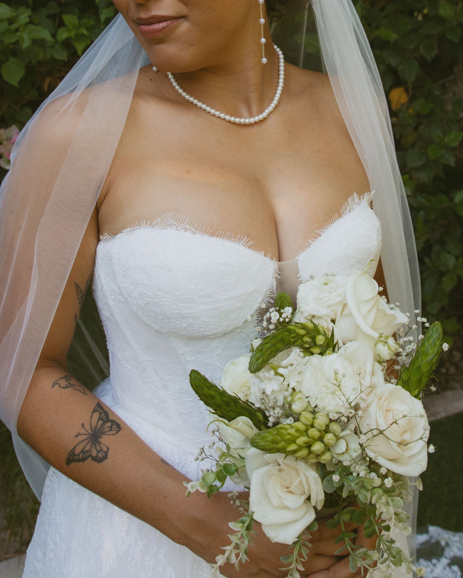Close-up of bride in strapless lace wedding dress holding white rose bouquet with pearl necklace and veil.